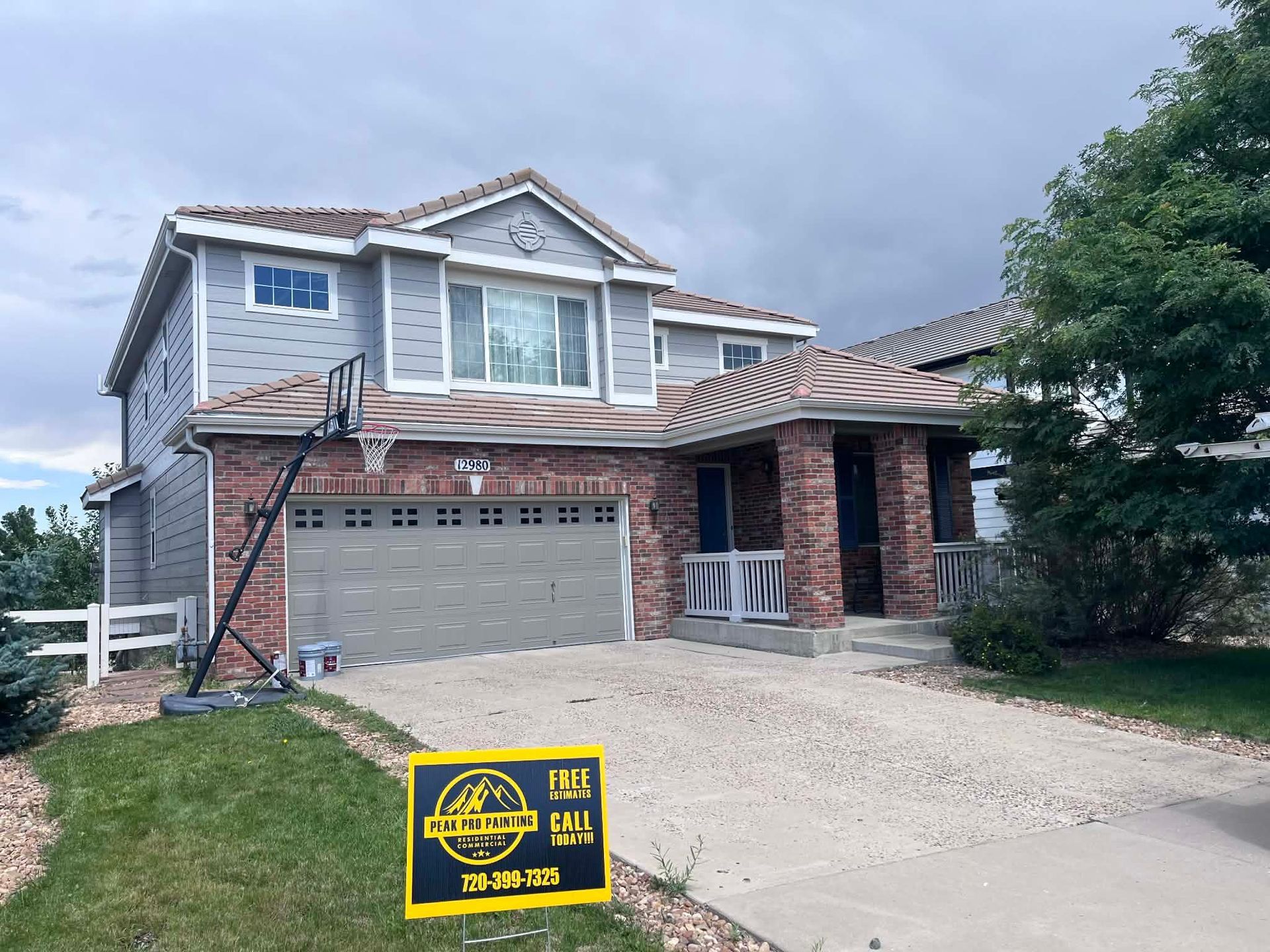 A two-story house with grey siding, brick facade, and a two-car garage, featuring a construction sign in the front yard.