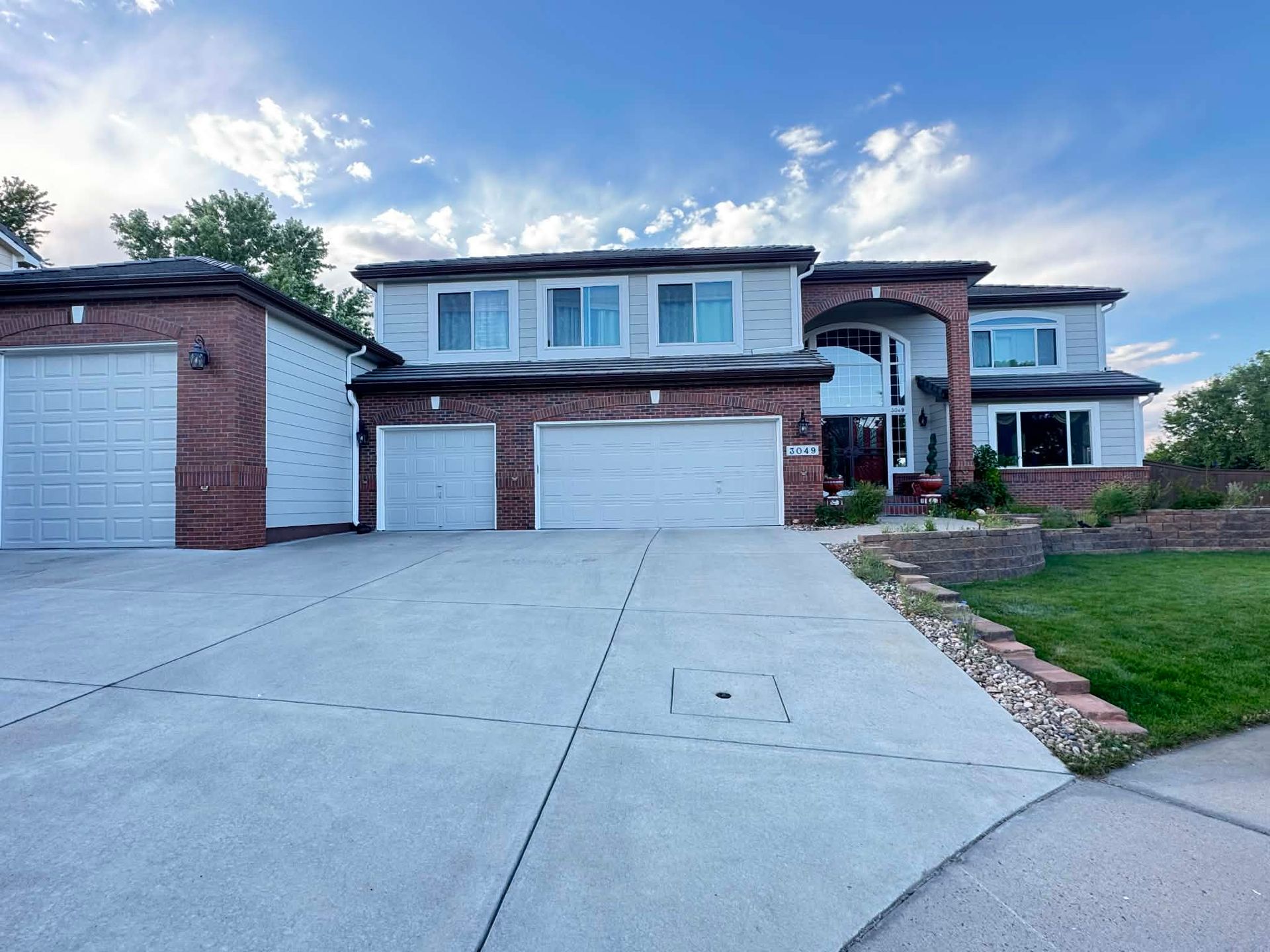 A two-story suburban home with a three-car garage, brick and siding exterior, and a paved driveway under a blue sky.