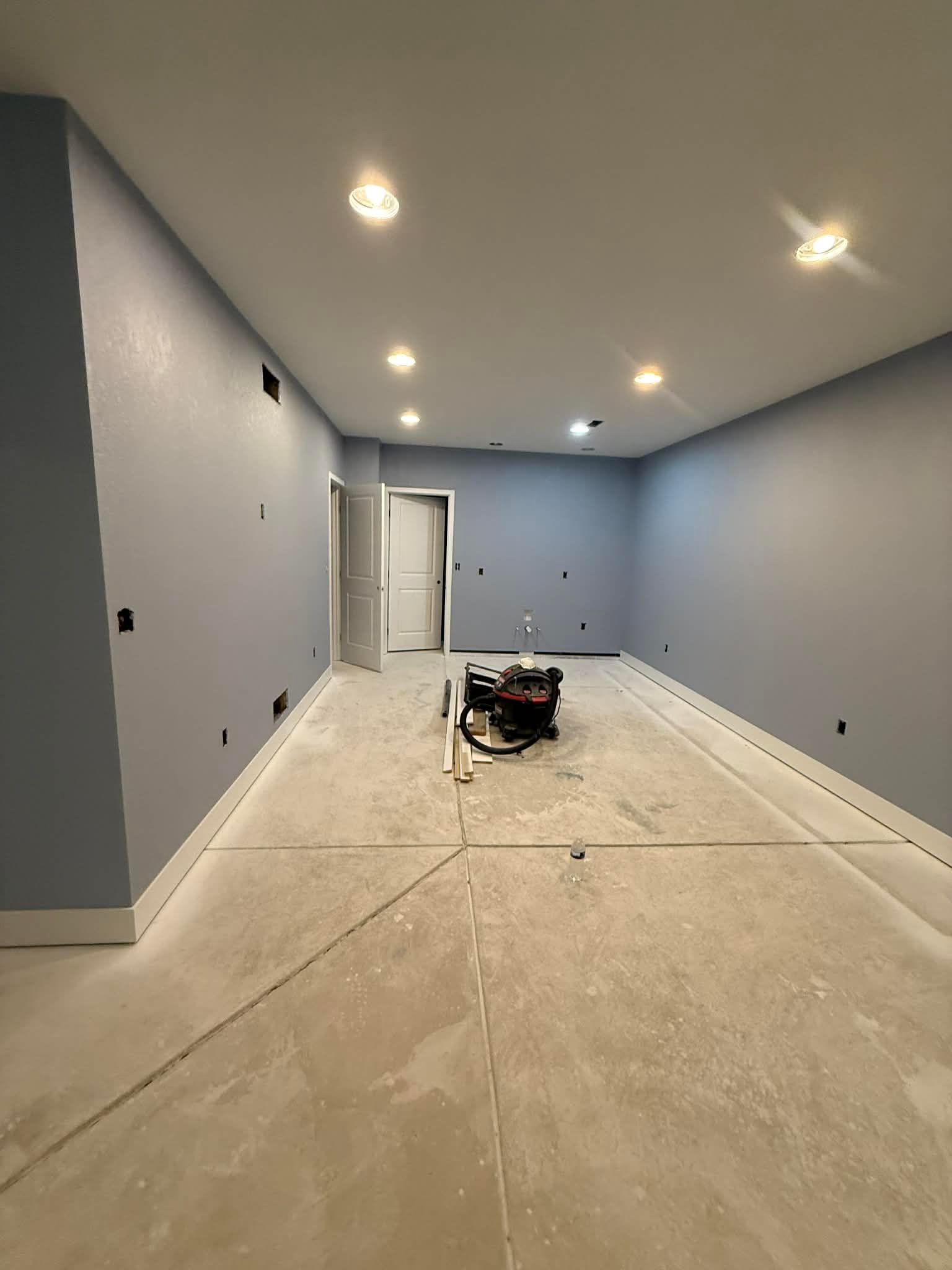 An empty room with blue-gray walls, white trim, recessed lighting, and a concrete floor under renovation.