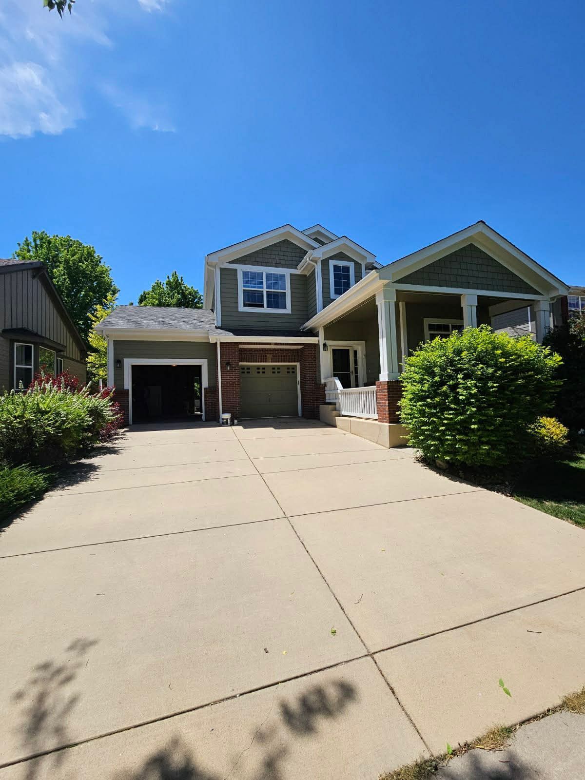 A two-story green house with a front porch, a concrete driveway, and an open garage, set under a clear blue sky.