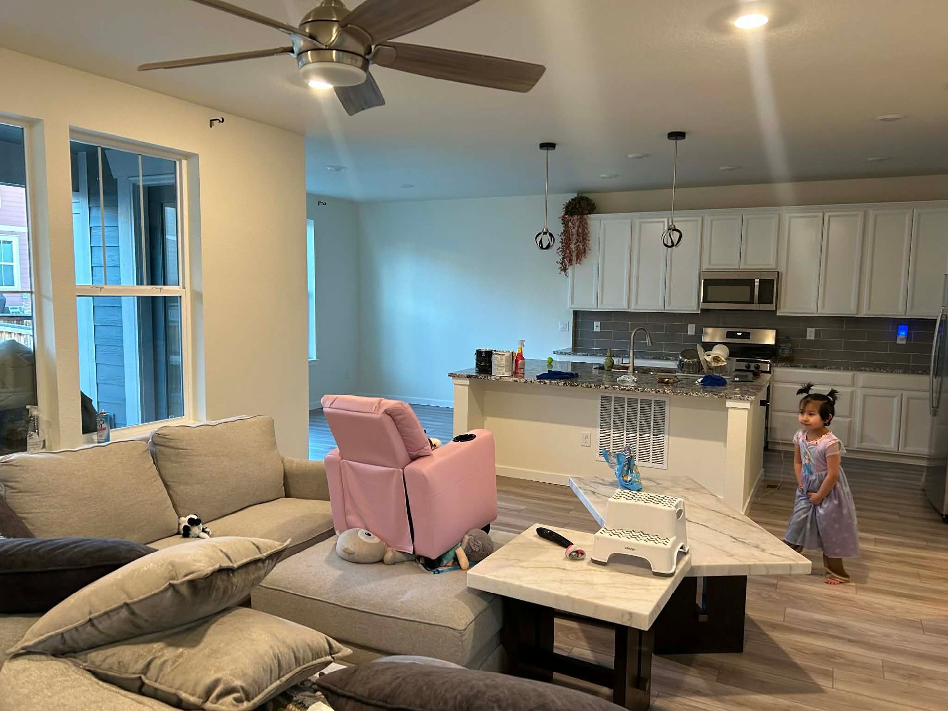 An open-concept room features a grey sectional, a pink chair, a marble coffee table, and a toddler standing in a kitchen.