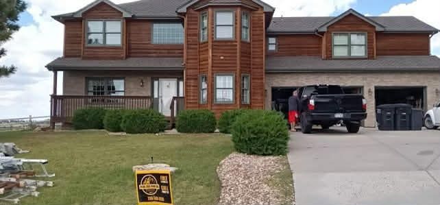 A two-story house with wood siding and a stone lower level, featuring a driveway with a dark truck parked in a garage.