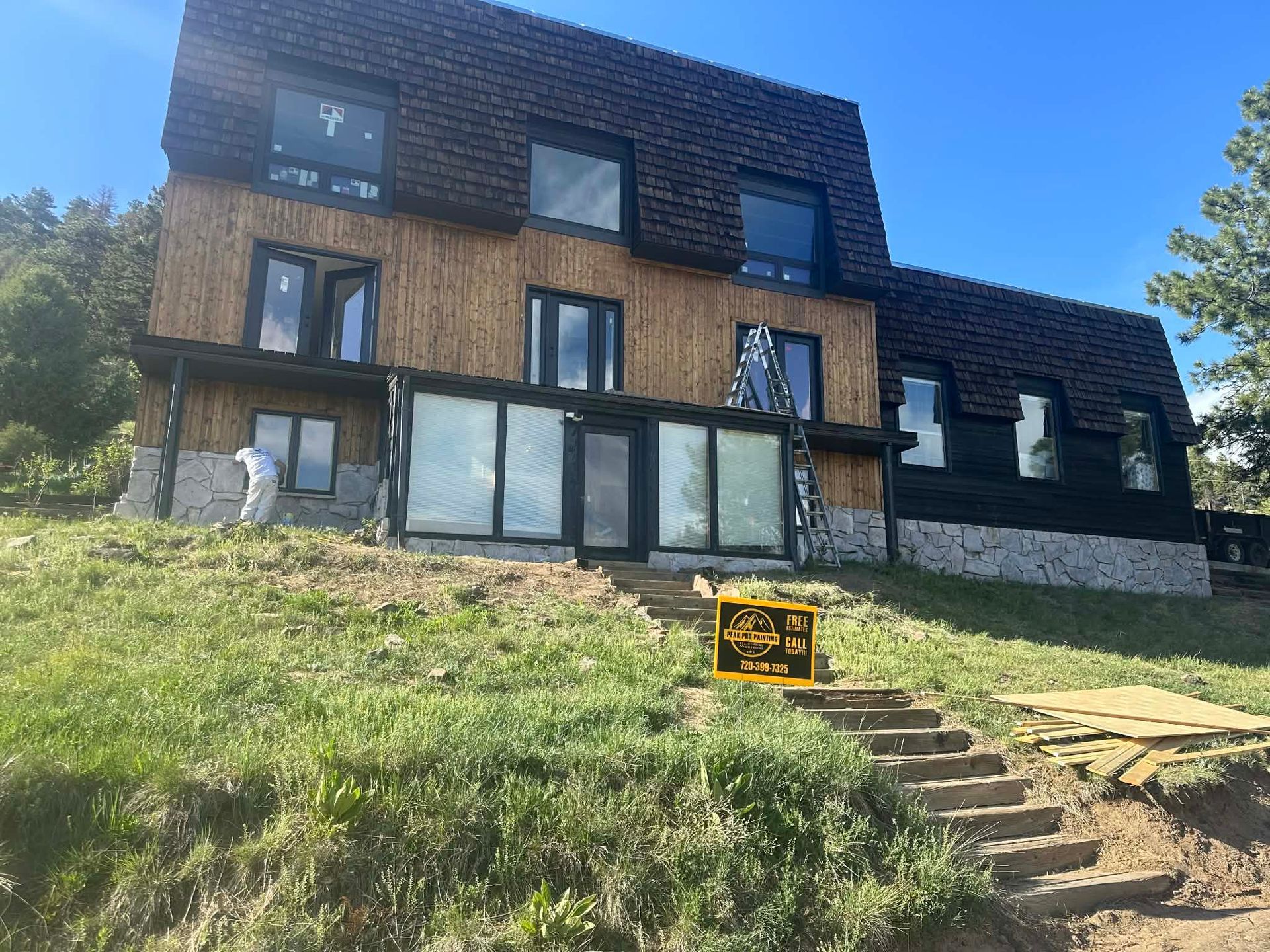 A house under construction with stone foundation, wooden siding, and a dark roof, viewed from a grassy hill with stairs.