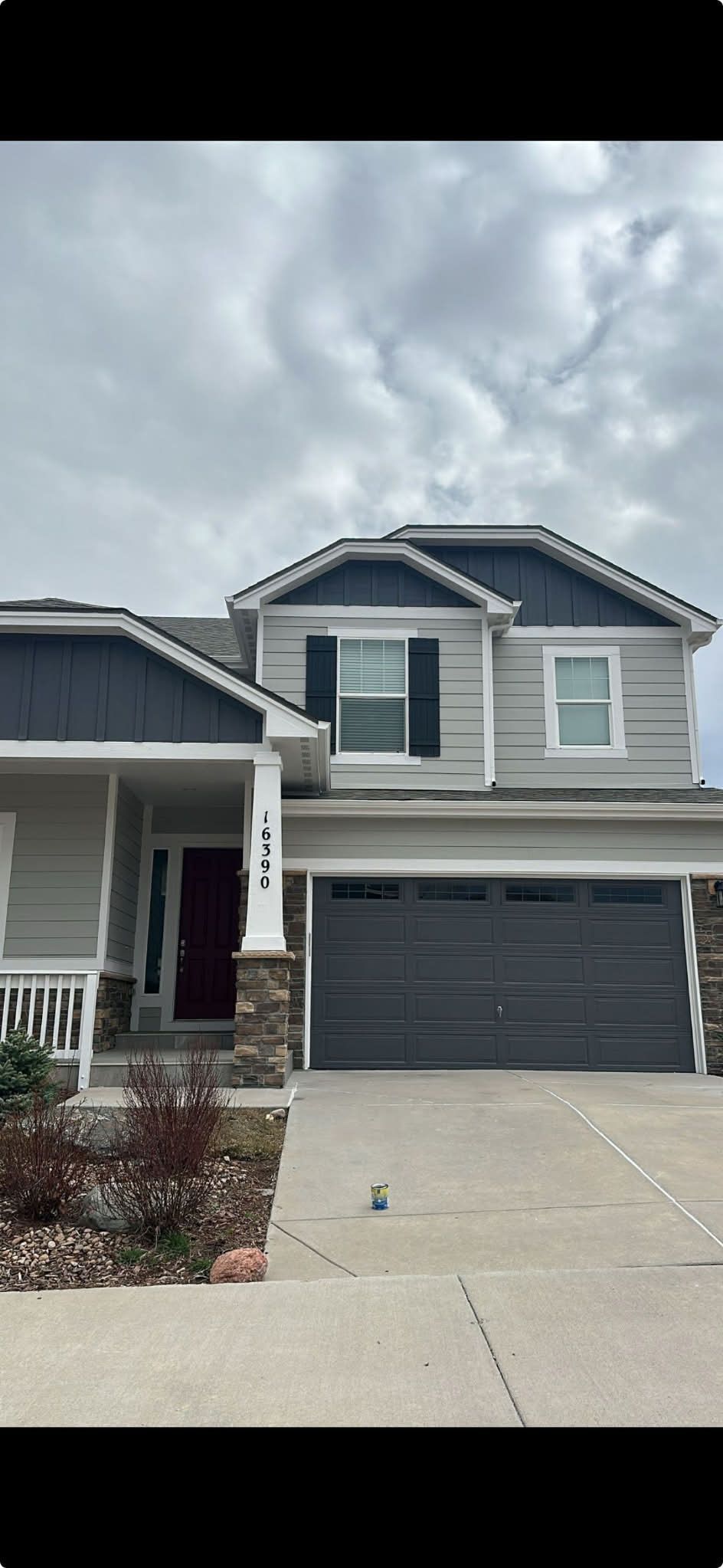 A two-story suburban house with grey siding, a dark blue garage, stone accents, and a covered front porch under a cloudy sky.