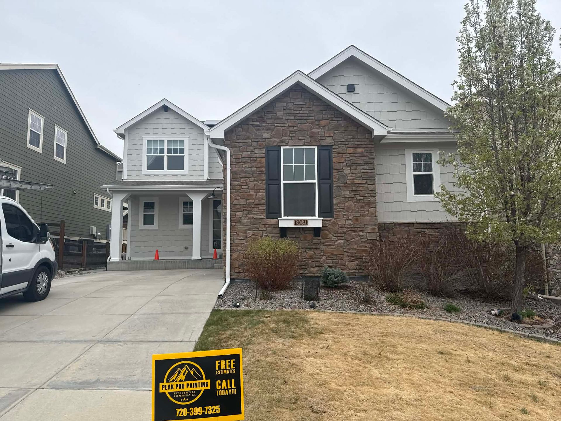 A two-story suburban house with stone and light grey siding, a concrete driveway, and a yellow sign in the front yard.