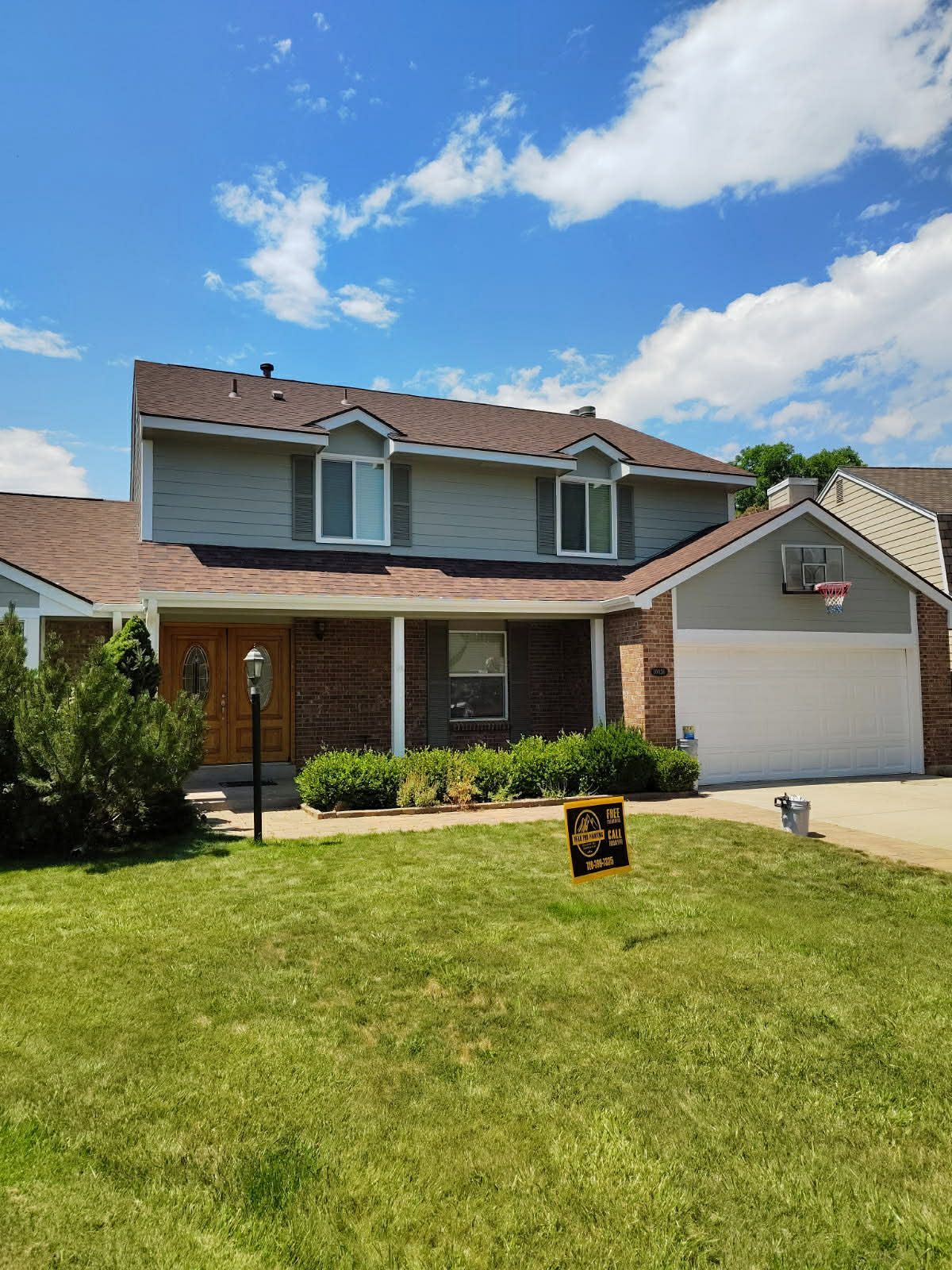 A two-story suburban house with a brick and gray siding exterior, a white garage, and a green front lawn under a blue sky.