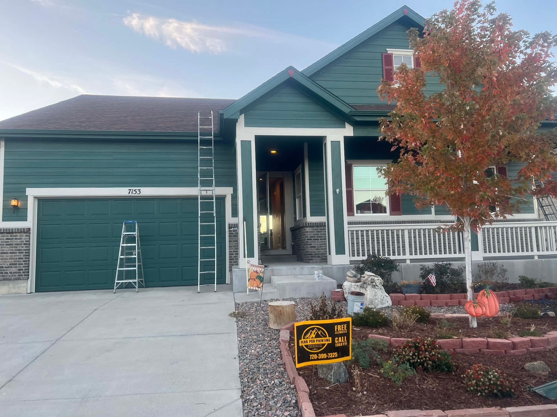 A dark green, single-story suburban house with a garage, front porch, and tall ladder against the roof during autumn.