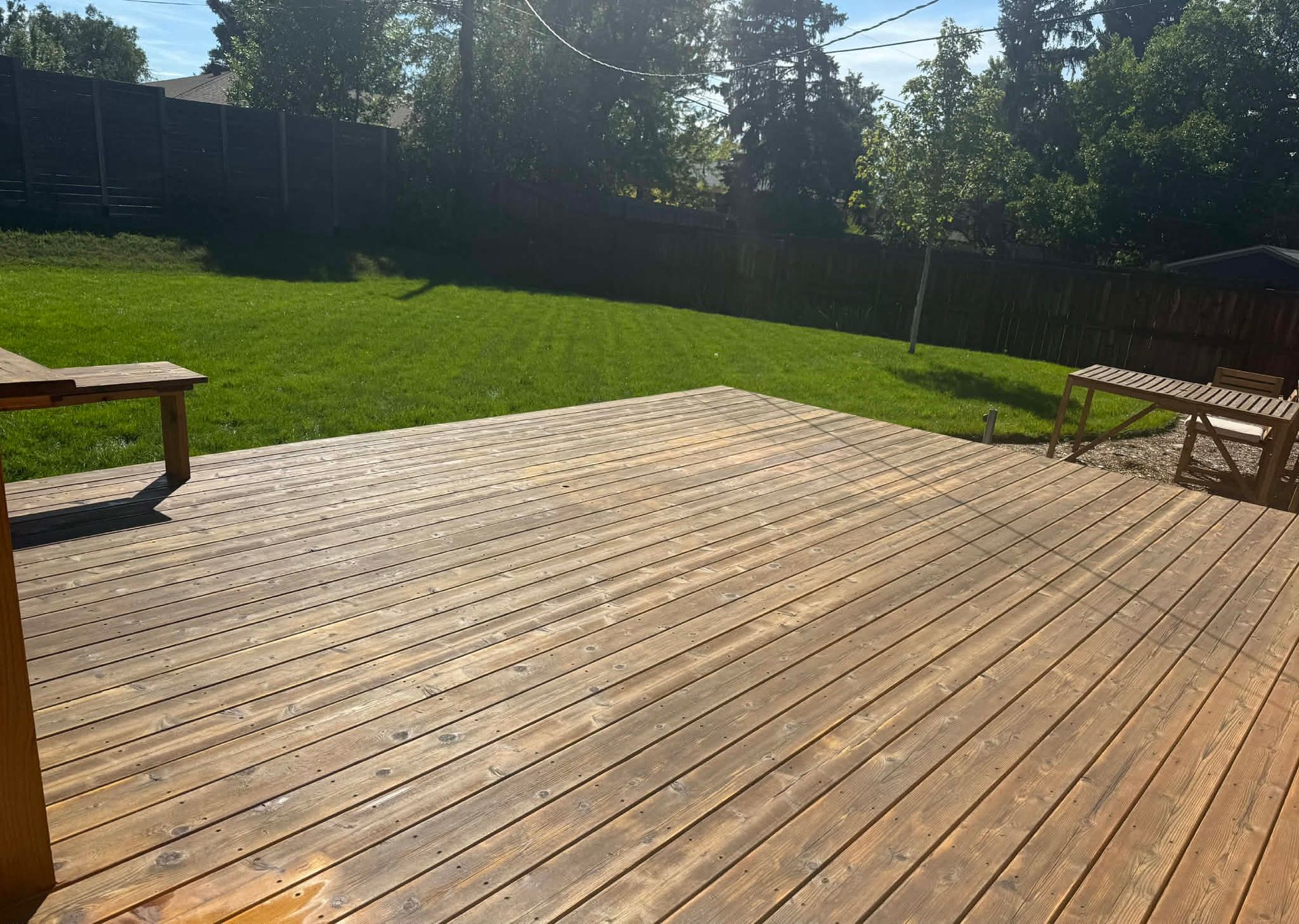 A wooden deck in a backyard with a large green lawn, surrounded by a fence and trees under a sunny sky.