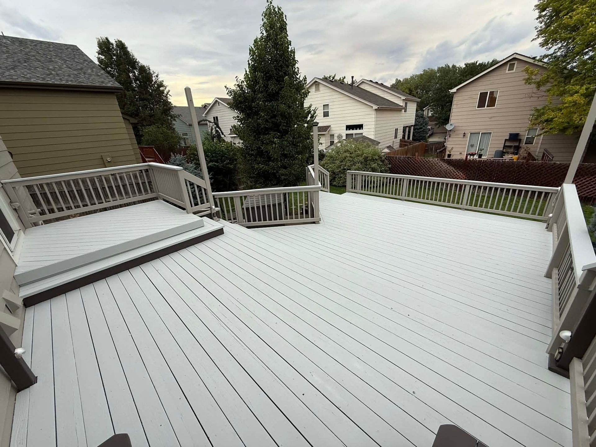 A large, light-gray wooden deck with railings, built-in seating, and steps, overlooking a suburban yard.
