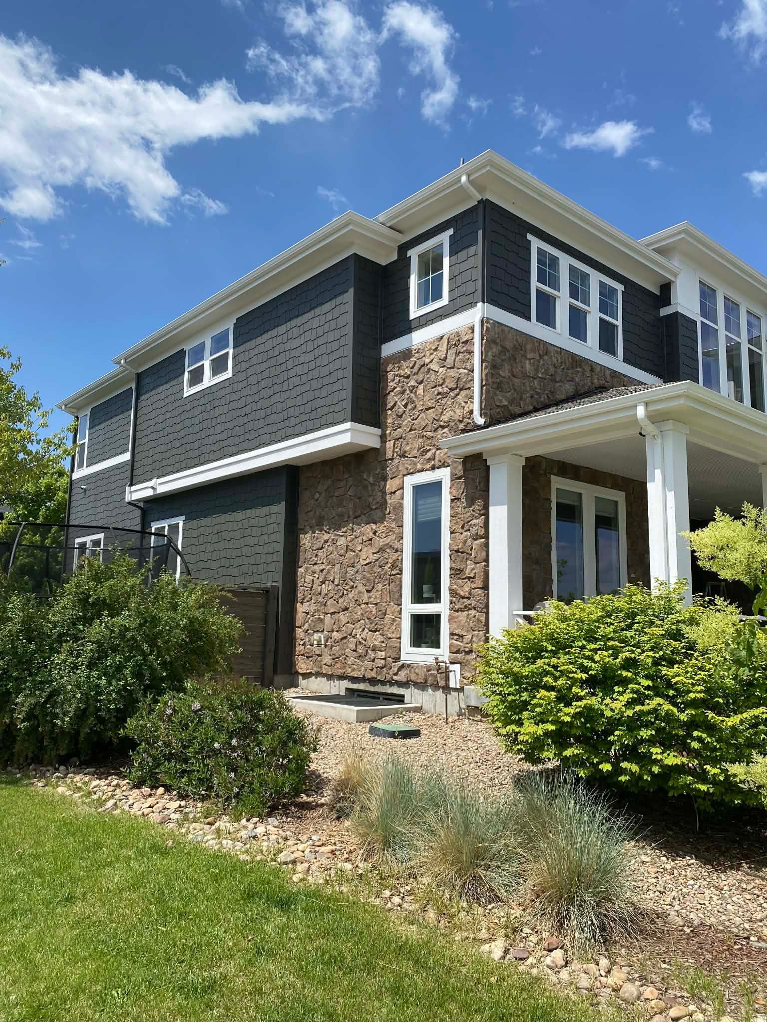 A two-story house with dark grey siding and stone veneer accents, viewed from a landscaped yard under a blue sky.