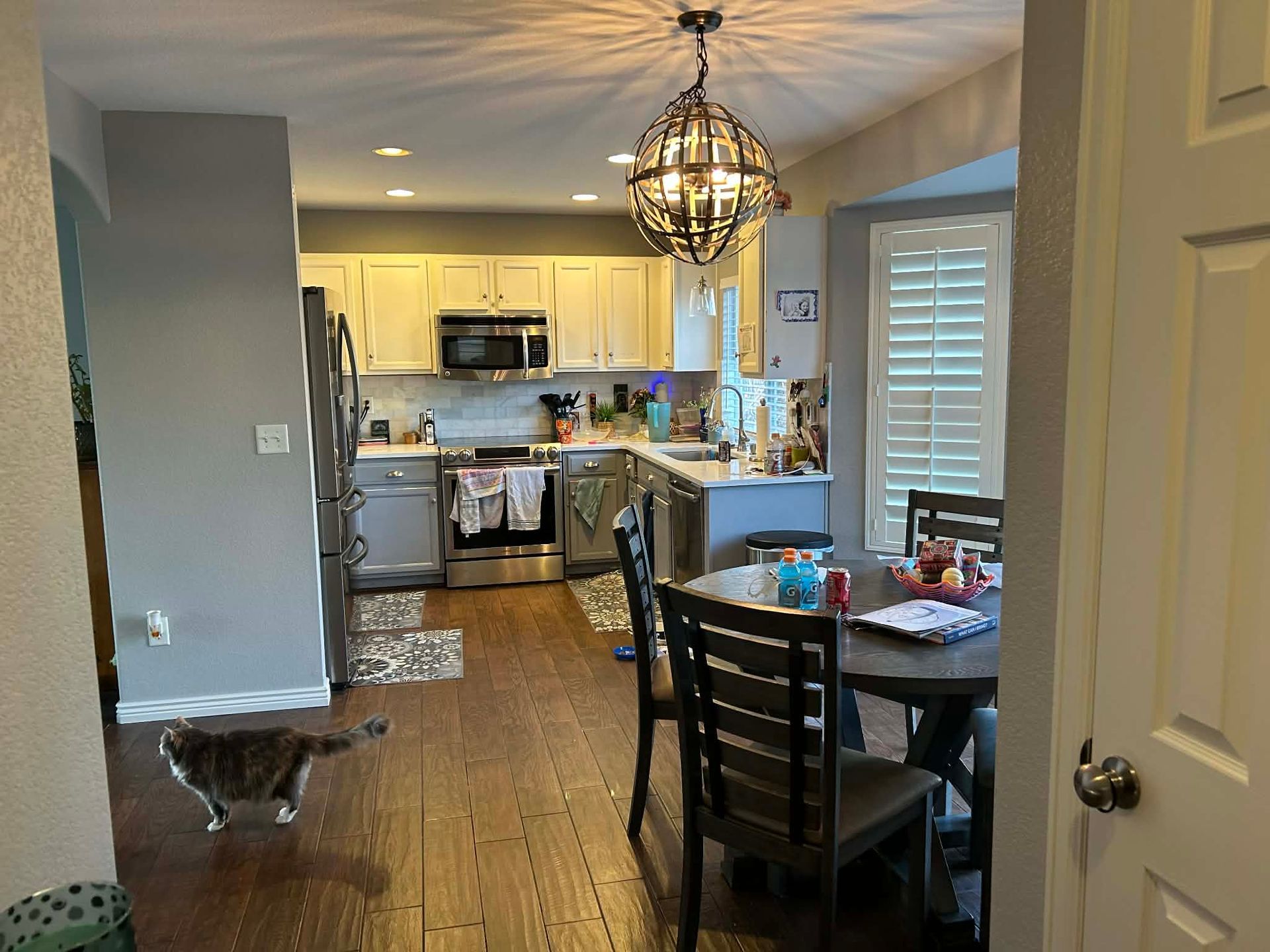 A grey cat walks across a wooden floor in a kitchen with white cabinets, stainless steel appliances, and a dining table.