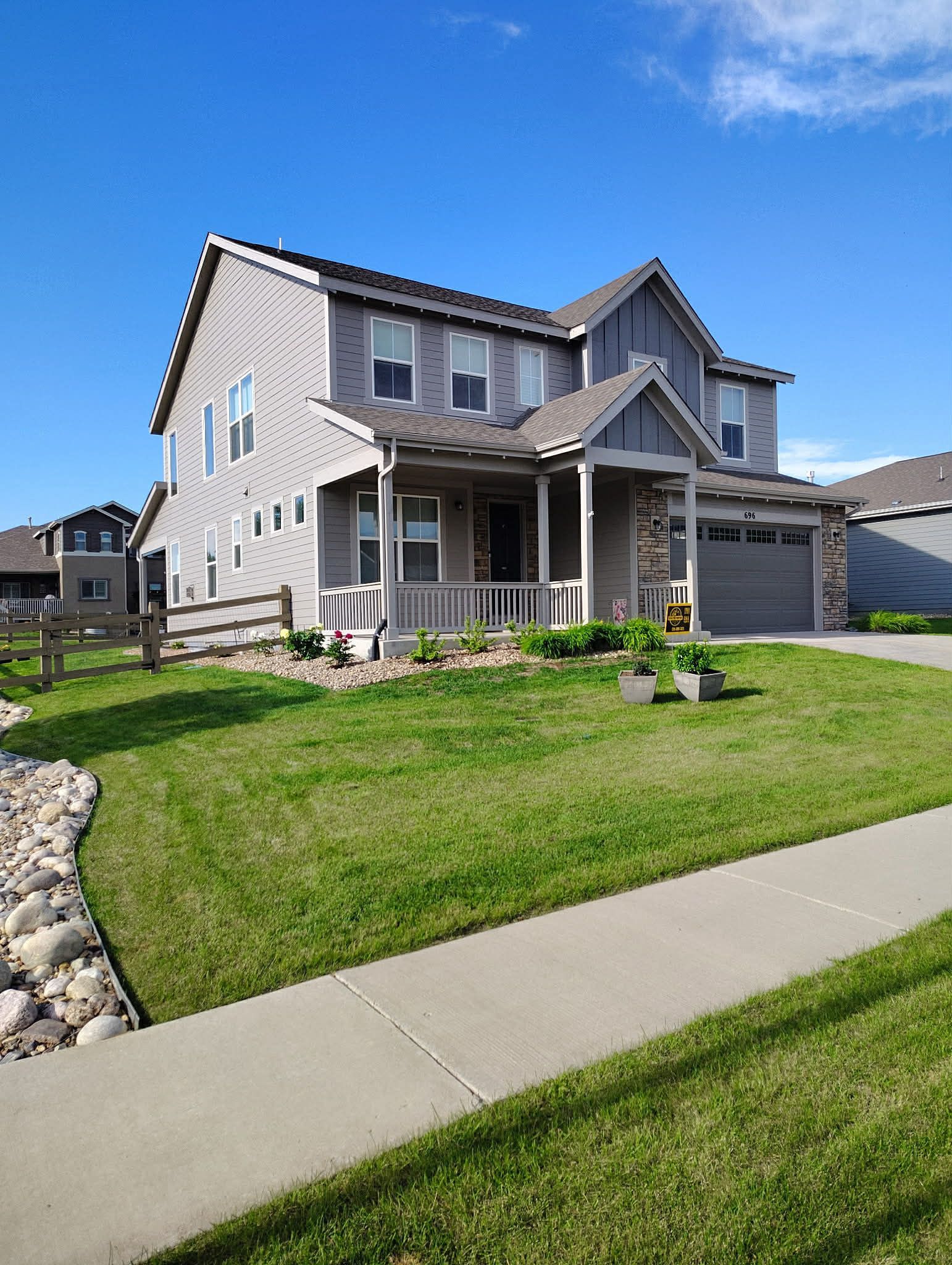 A two-story suburban house with light gray siding, stone accents, and a front porch under a bright blue sky.