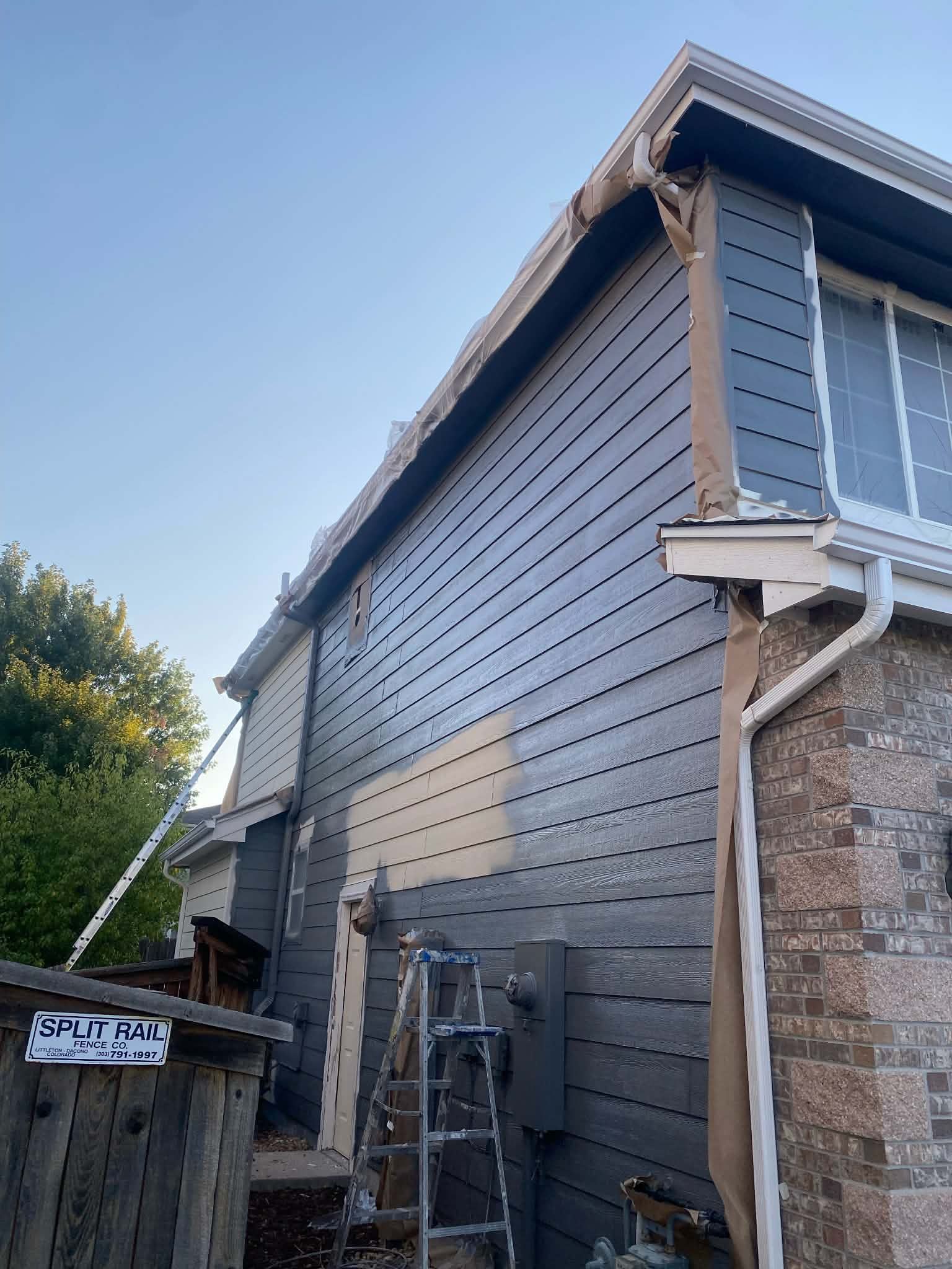 A side view of a house undergoing siding installation, with blue panels partially covering the wall and a ladder nearby.