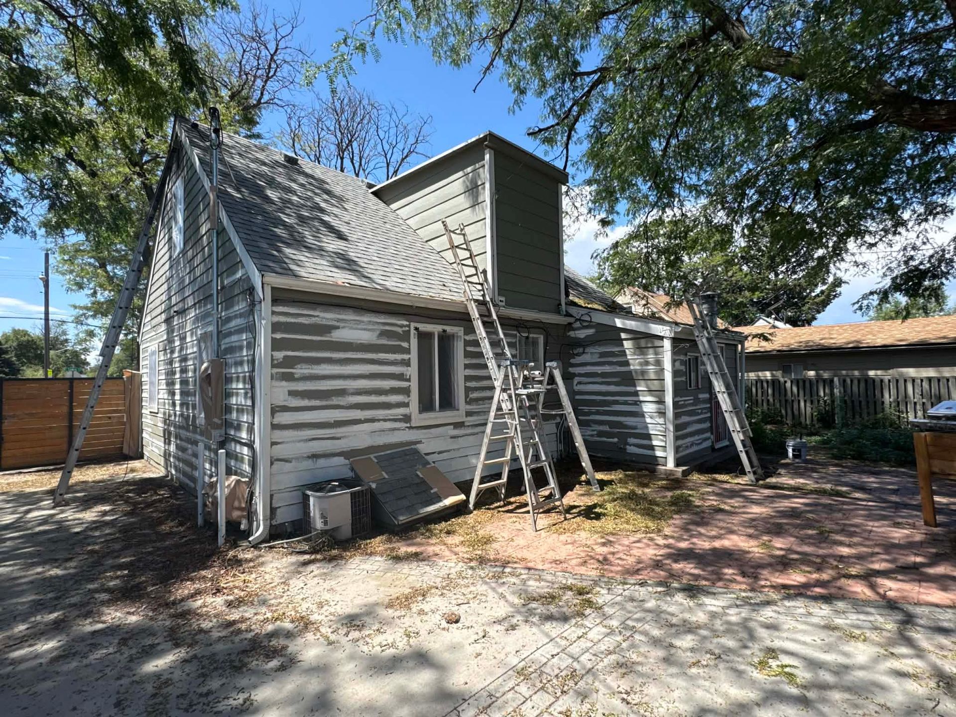 A two-story house with peeling white paint and gray siding, undergoing exterior repairs with ladders against the walls.