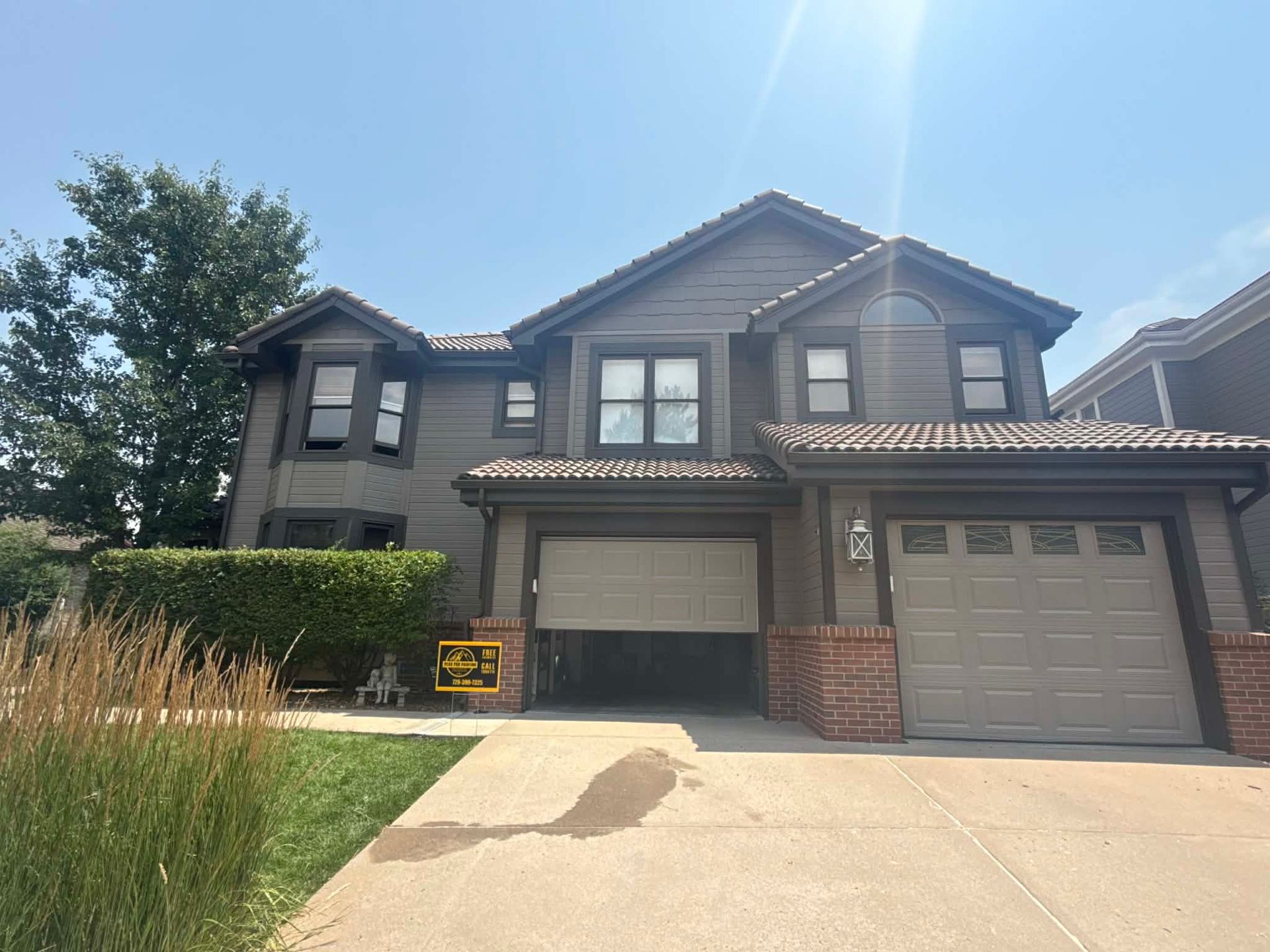A two-story, gray suburban house with a brown roof, two garage doors, and a front yard with grass and tall decorative grass.