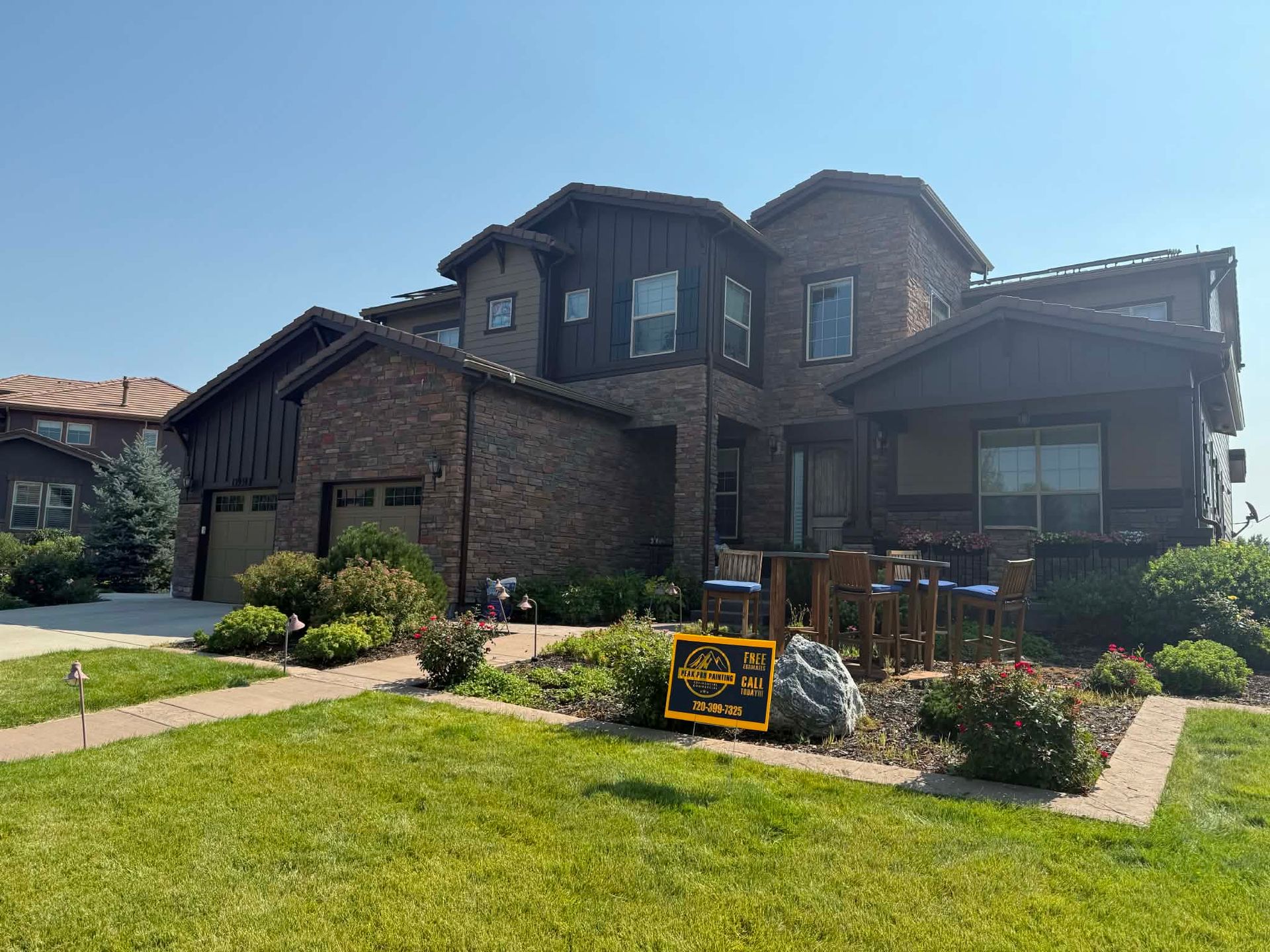 A two-story brown brick house with a garage, dark trim, and a small sign in the front yard garden.