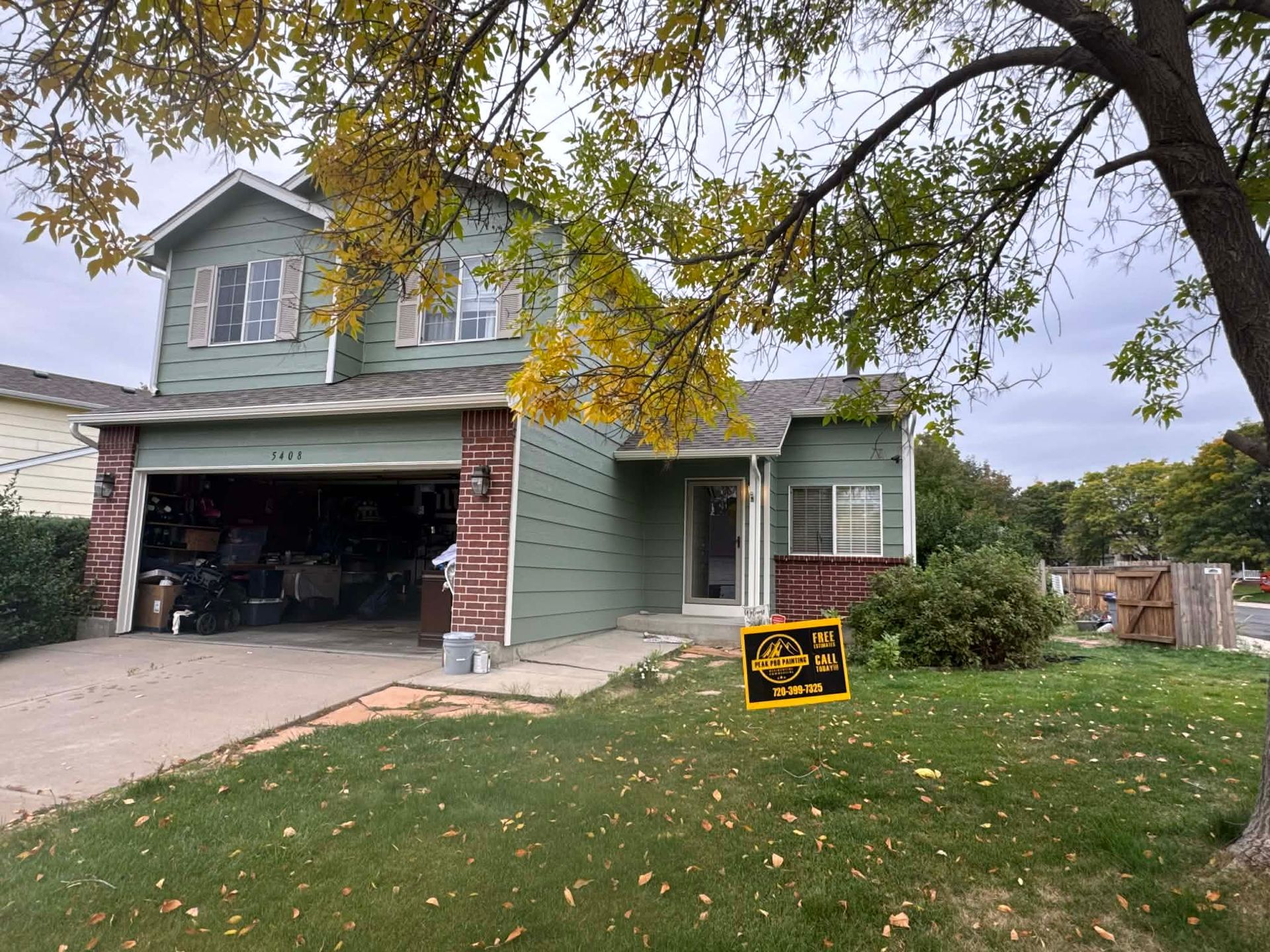 A sage green, two-story house with a garage, brick accents, and a front yard with a yellow sign under a large autumn tree.