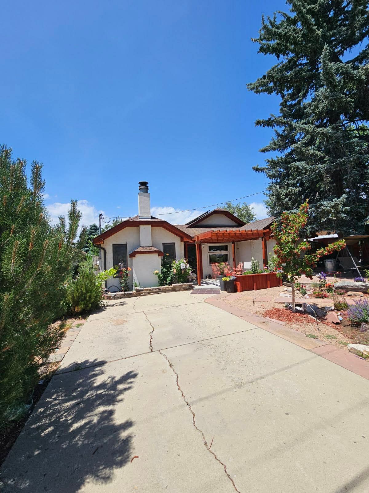 A white, single-story house with a prominent chimney, red-brown trim, and a wooden pergola over a front seating area.