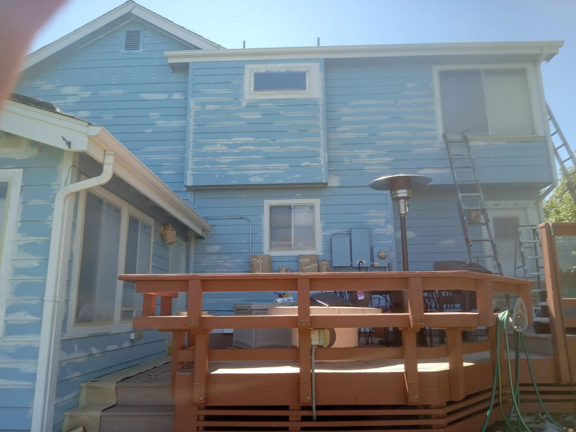 A light blue house exterior with peeling paint, featuring a brown wooden deck, windows, and an extension ladder.