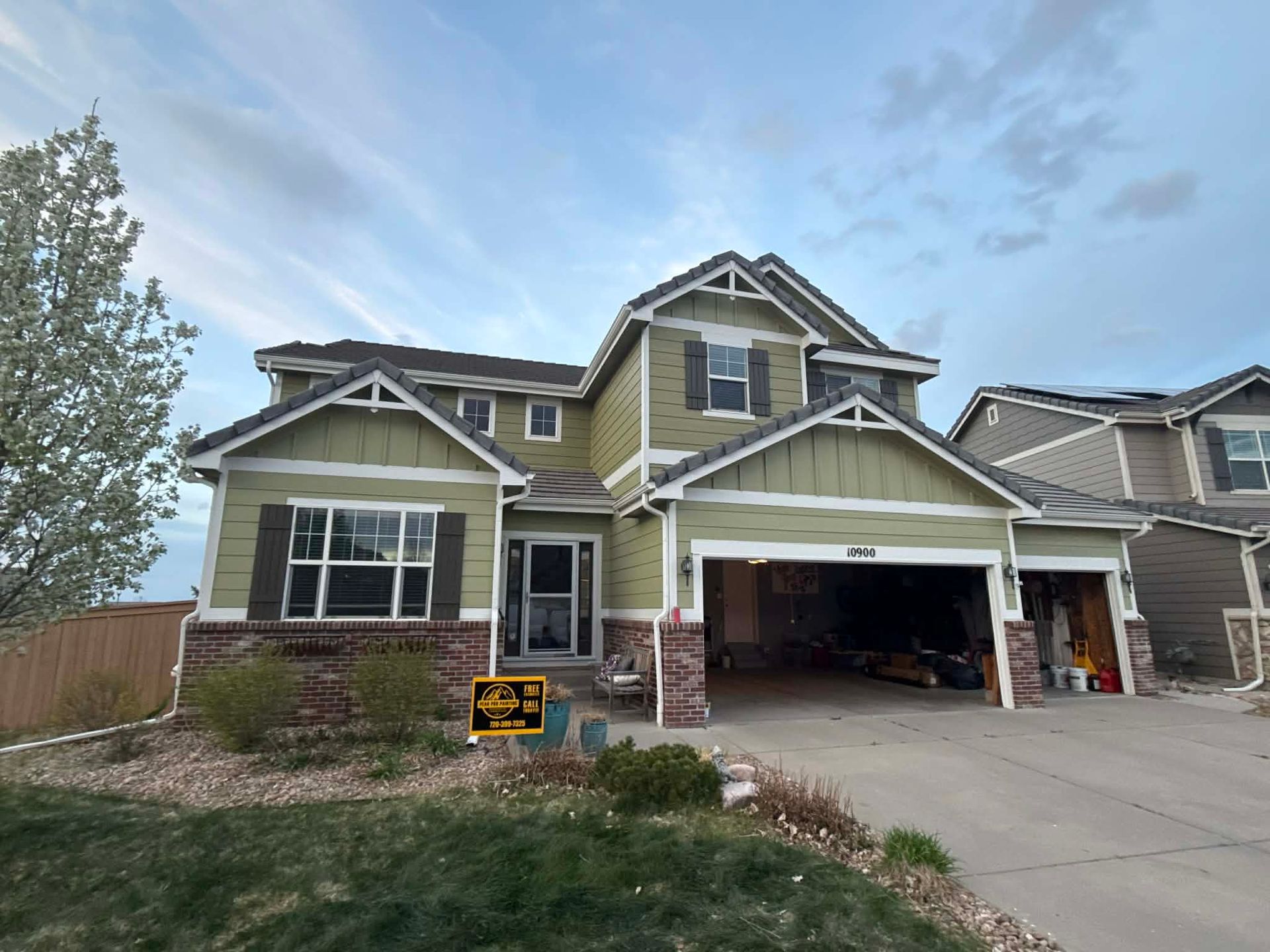 A two-story sage green house with a stone base, a double garage, and a large front window under an evening sky.