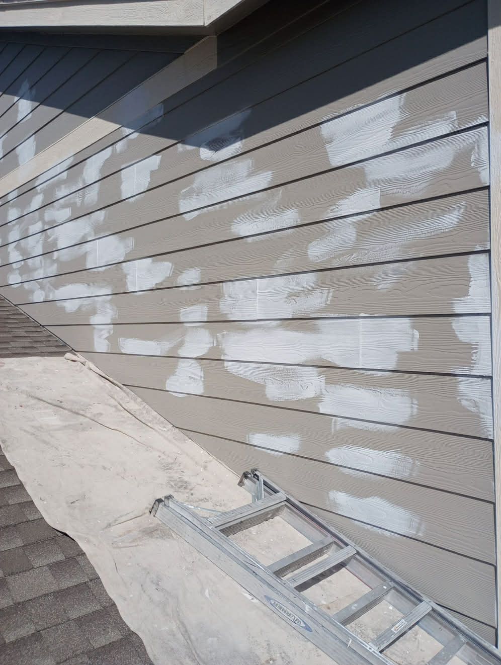 Tan horizontal siding on a house exterior featuring several patches of white primer or filler, with a ladder below.