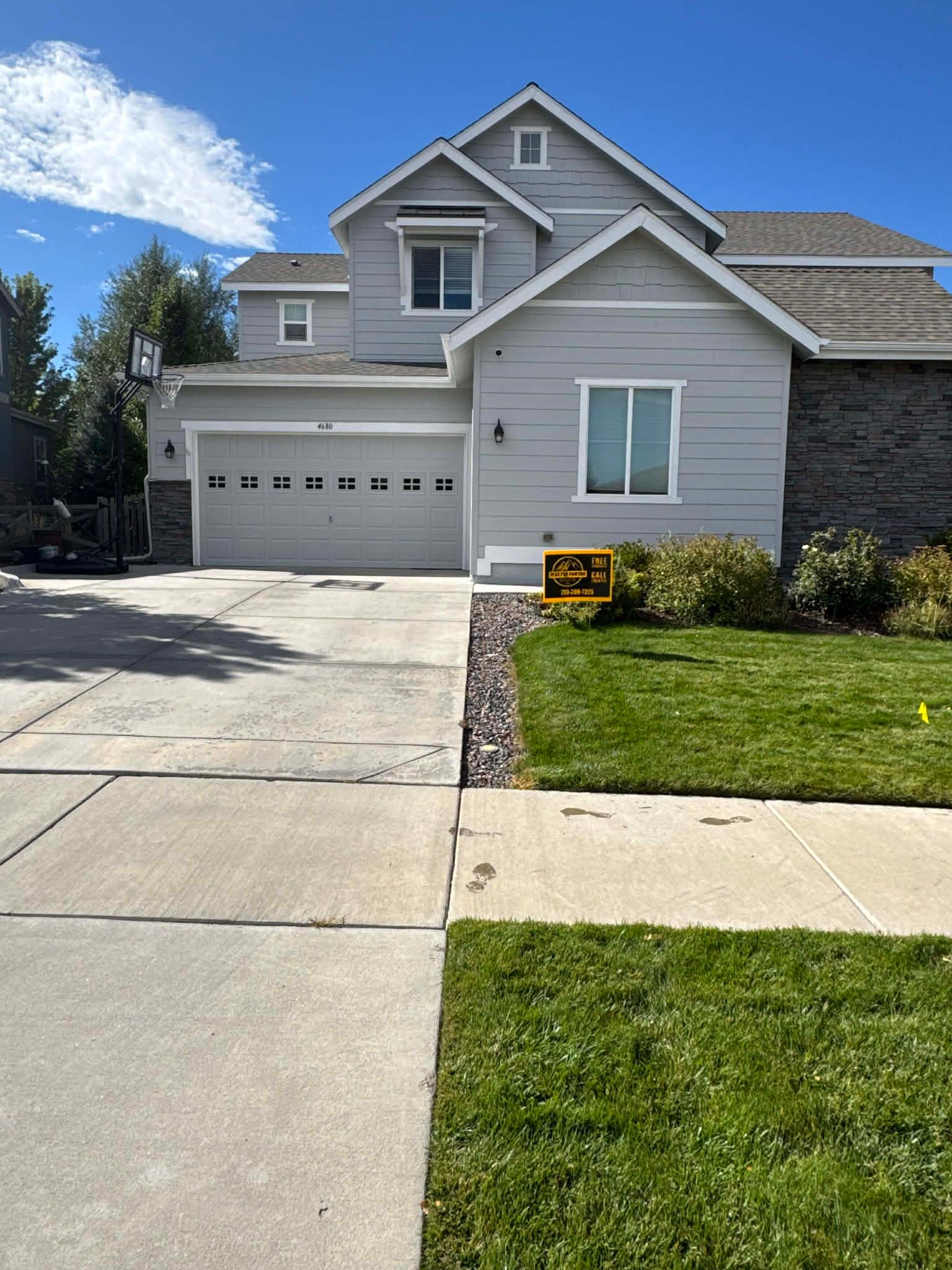 A gray, two-story suburban house with a two-car garage, a stone facade, and a well-maintained front lawn under a blue sky.