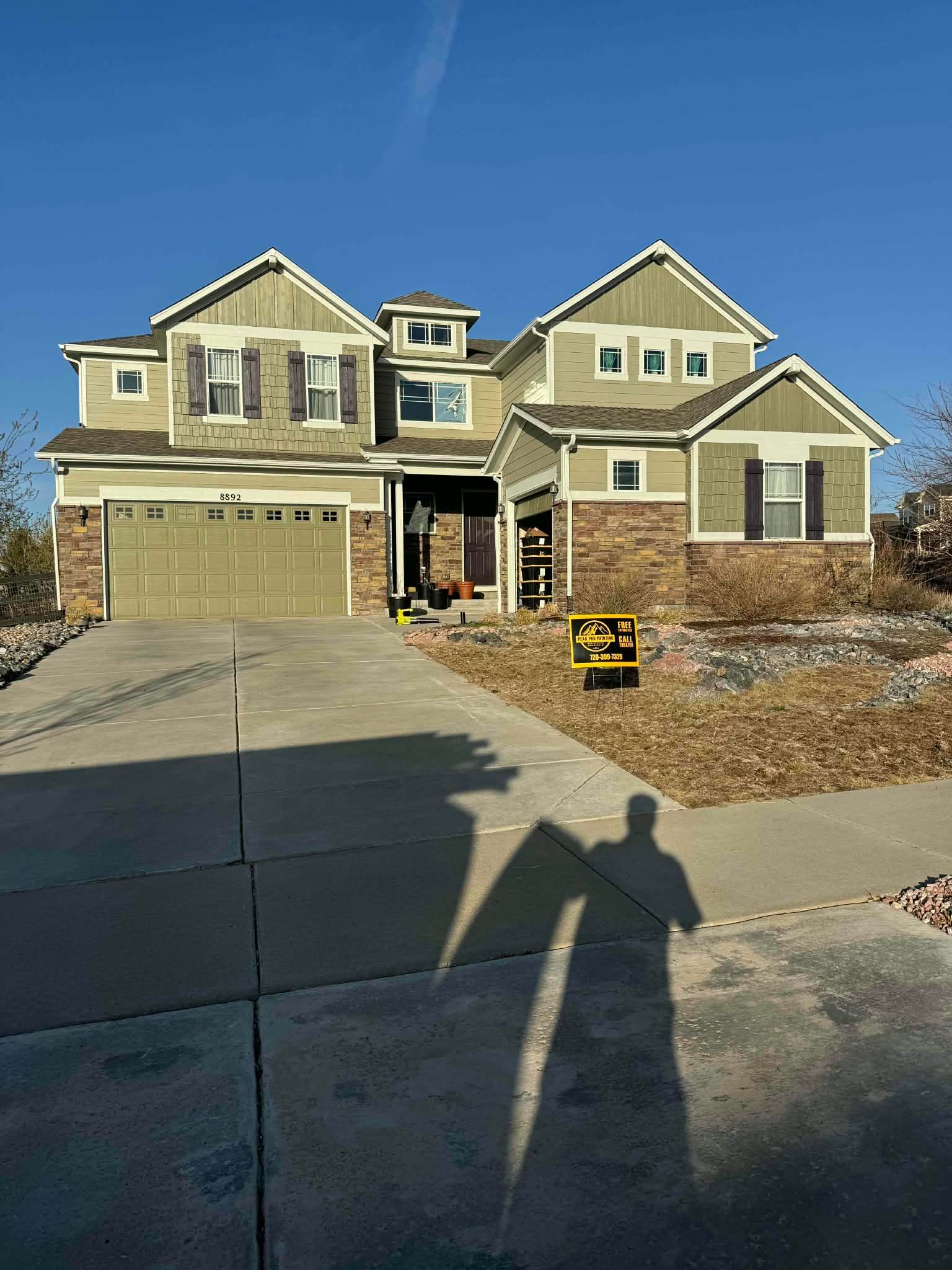 A tan two-story house with stone accents, a garage, and a shadow of a person in the foreground on a sunny day.