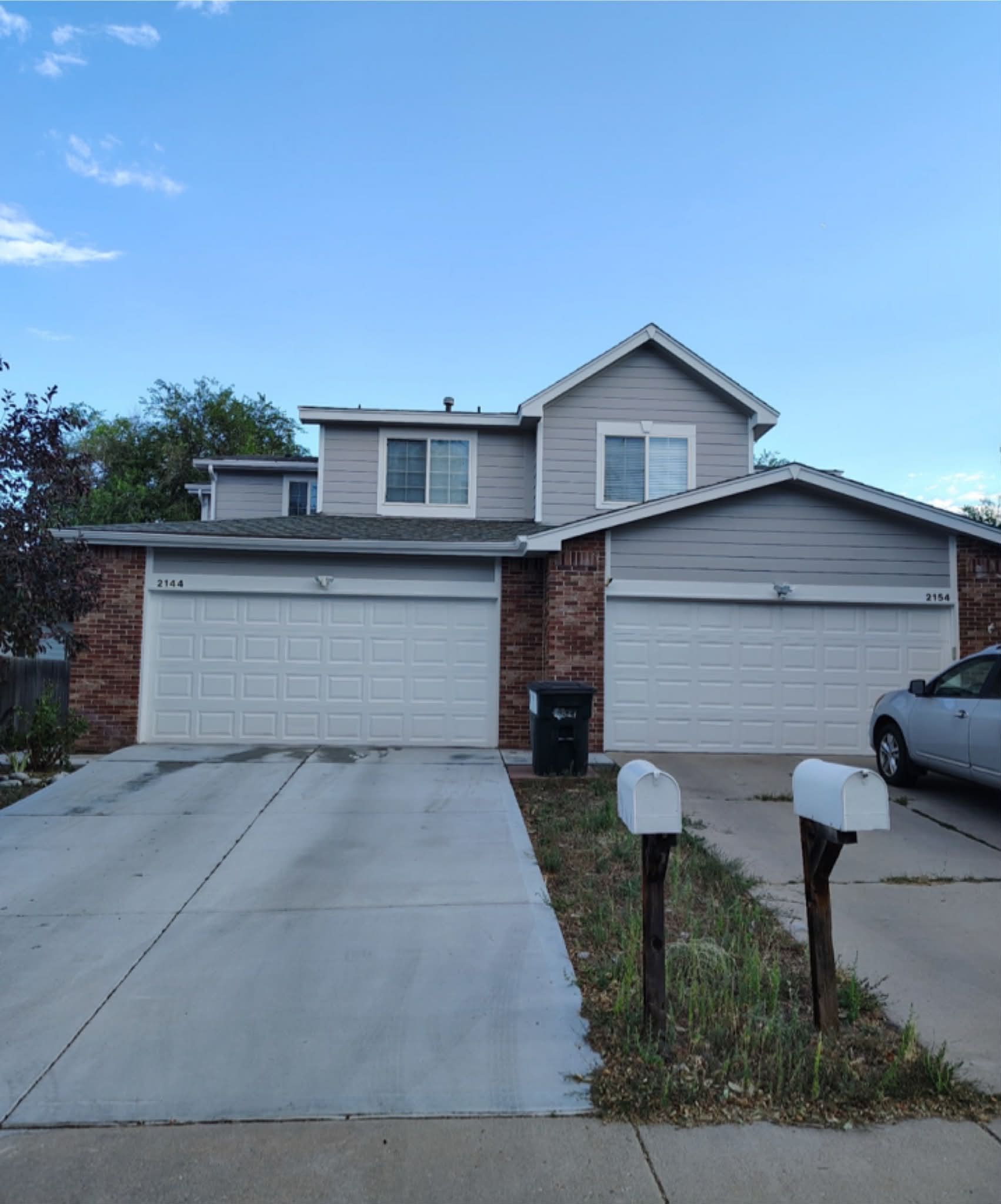 Two-story duplex with gray siding and brick accents, two white garage doors, and two mailboxes in the front yard.