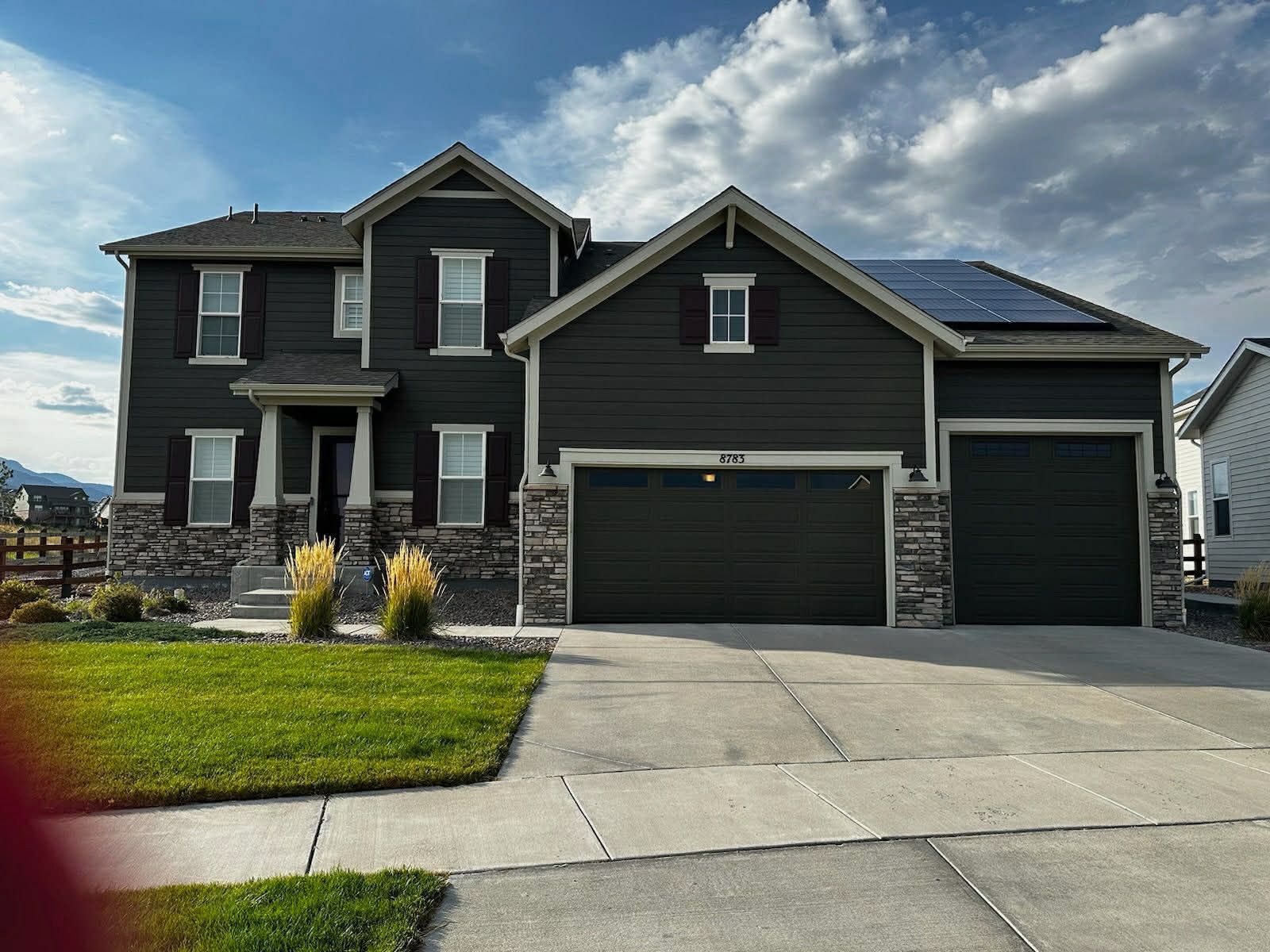 A two-story dark gray house with stone siding, a three-car garage, and rooftop solar panels under a partly cloudy sky.