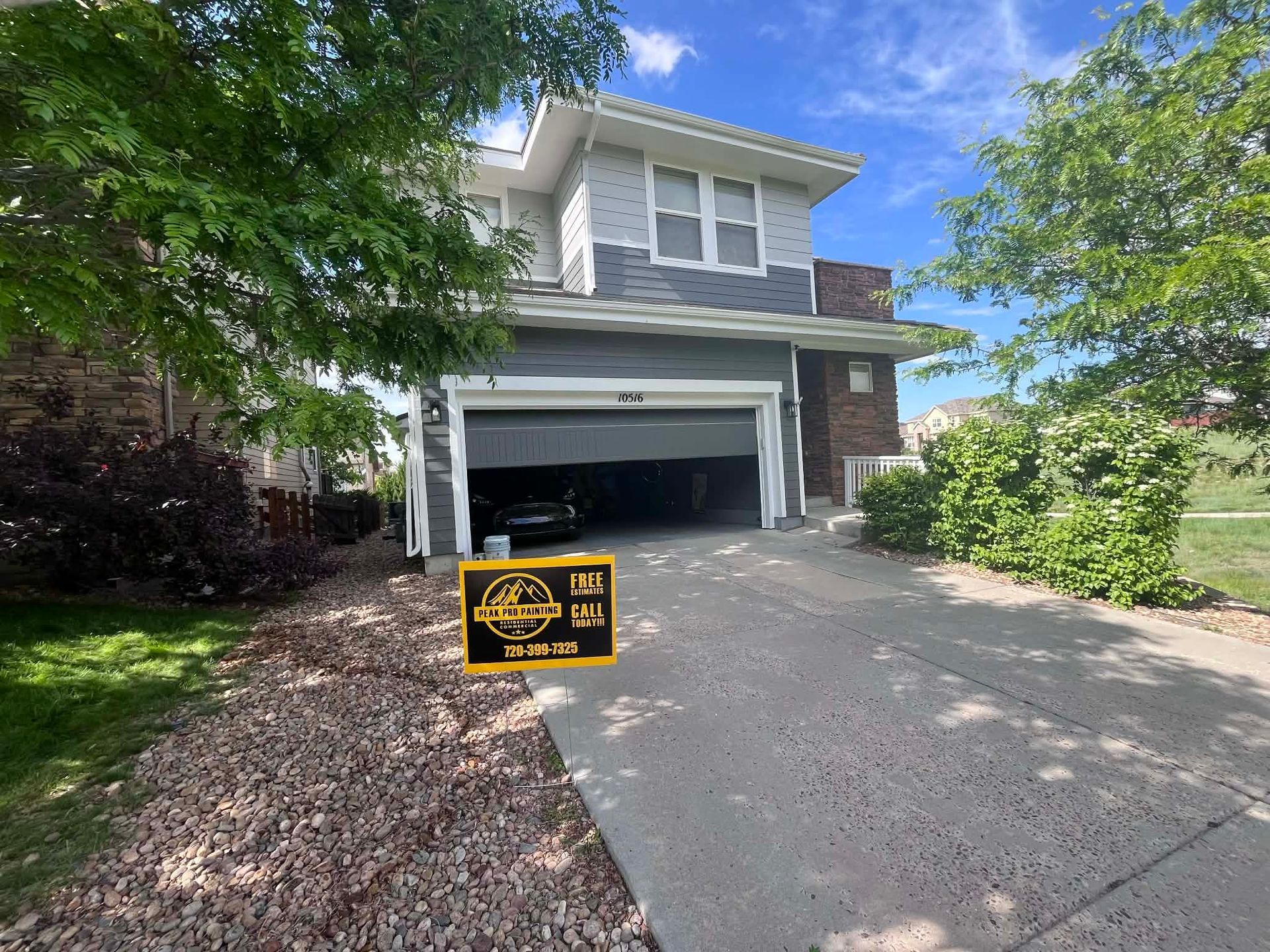 A two-story gray house with a partially open garage, a concrete driveway, and a yellow sign sitting on the front lawn.
