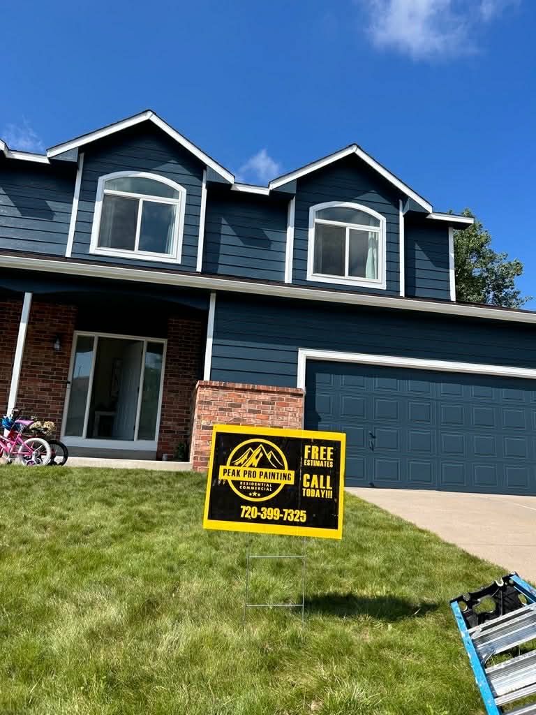 A blue, two-story house with a brick entryway and a promotional sign in the lawn on a sunny day.