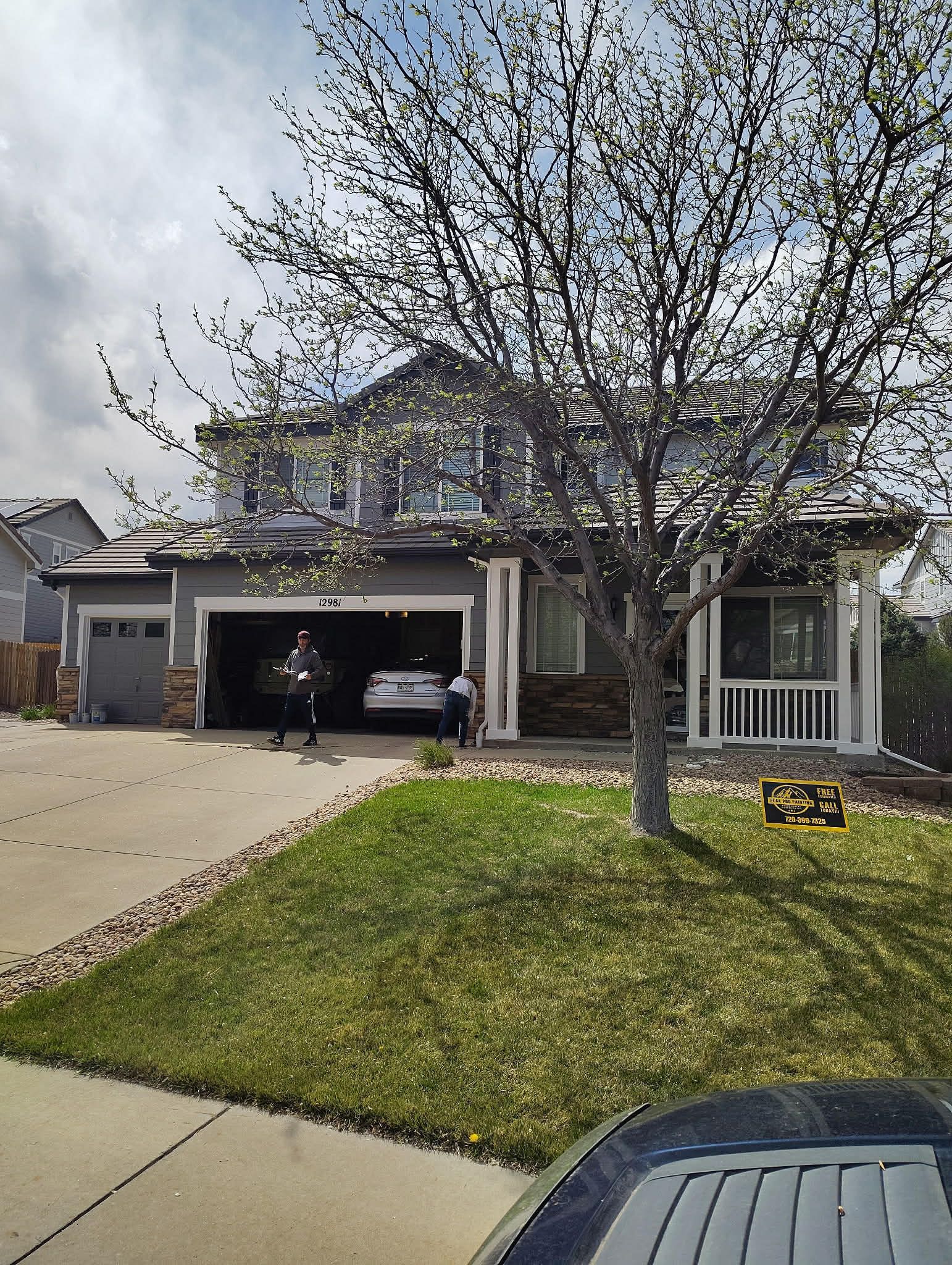 A gray, two-story house with a three-car garage, a front lawn with a large tree, and a driveway under a cloudy sky.