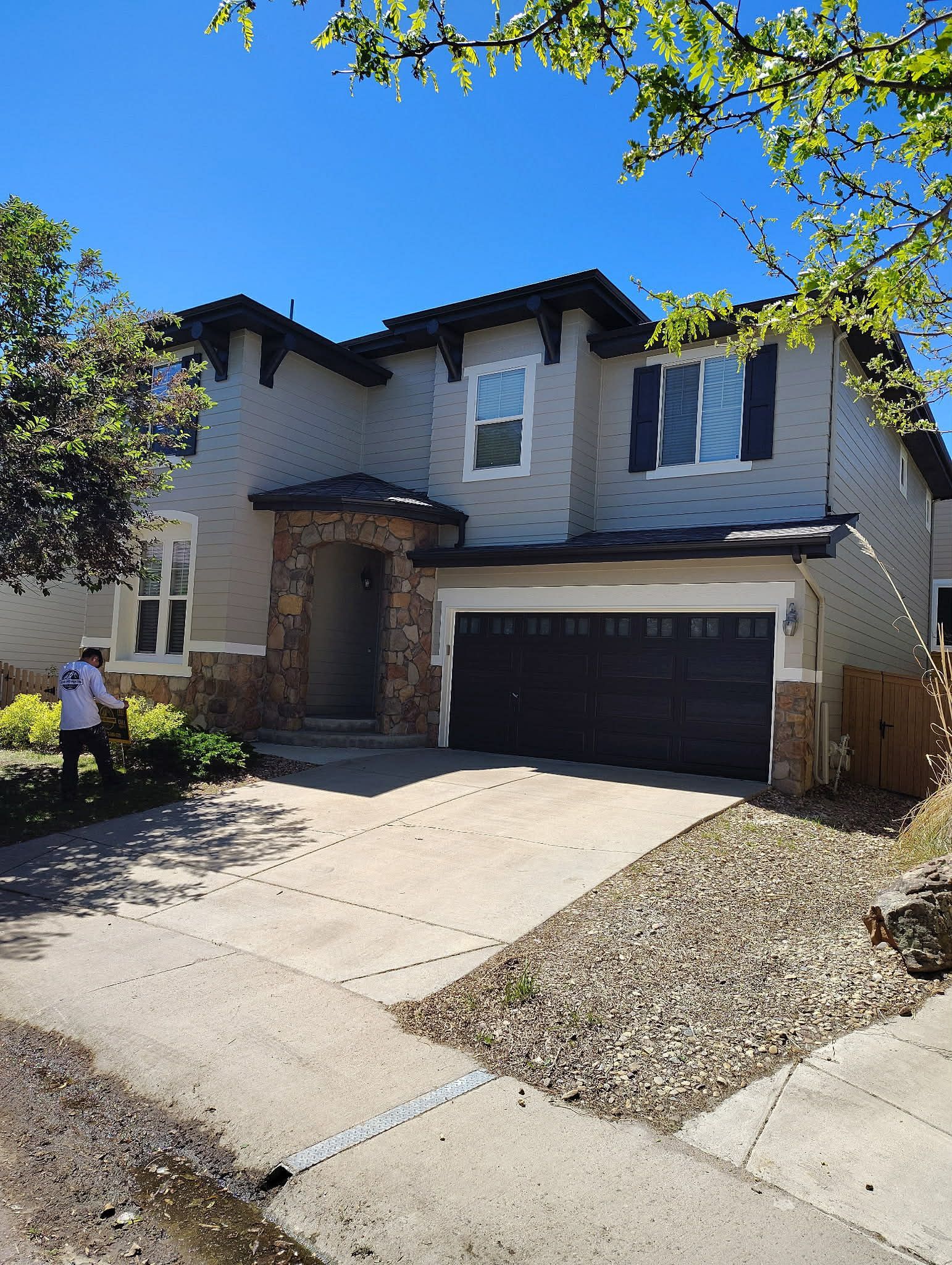 A two-story stucco house with a dark garage, stone accents, and a person standing in the front yard.