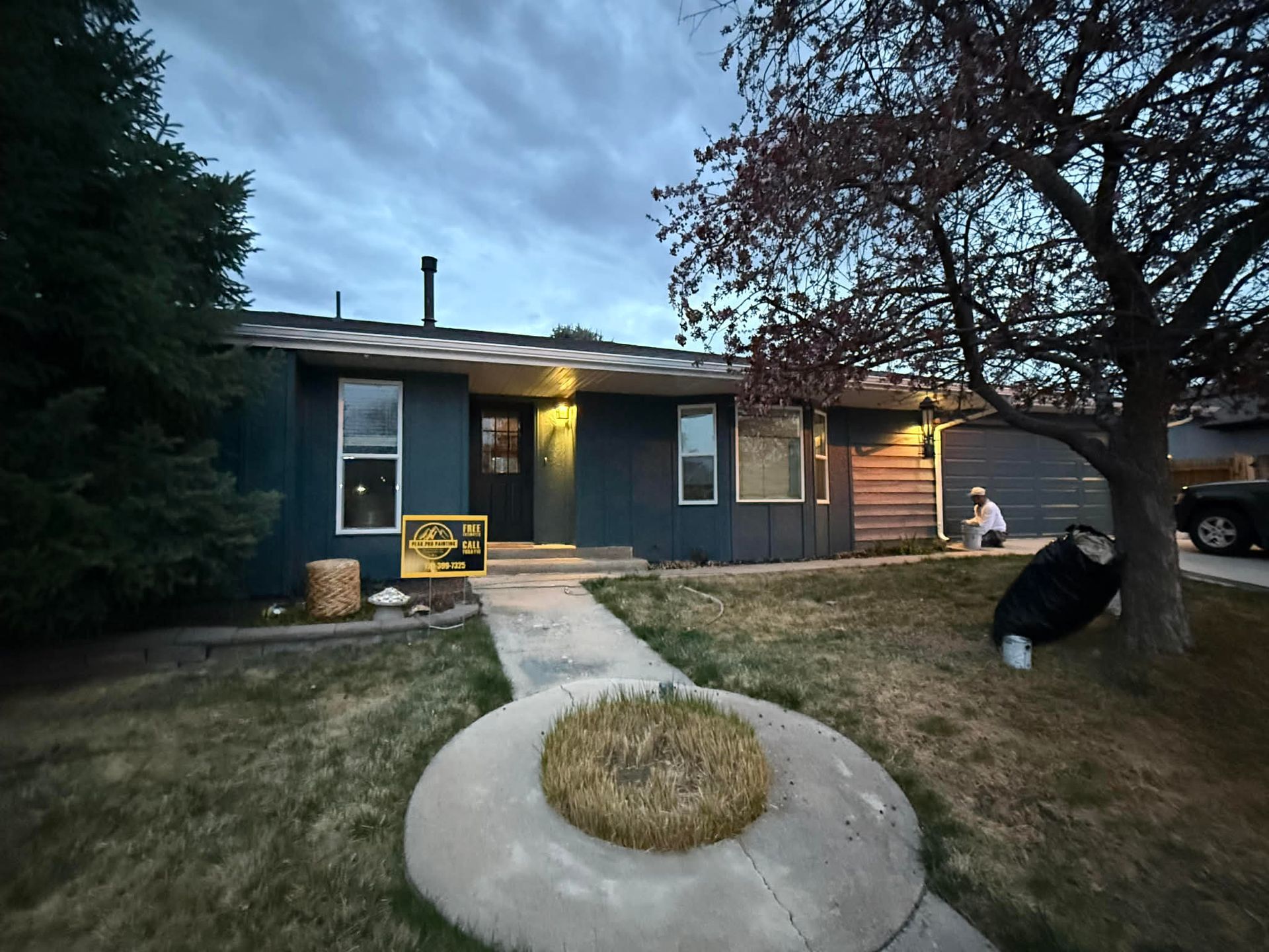 A dark blue single-story home at dusk with a circular planter, a sign near the front door, and a tree on the right lawn.