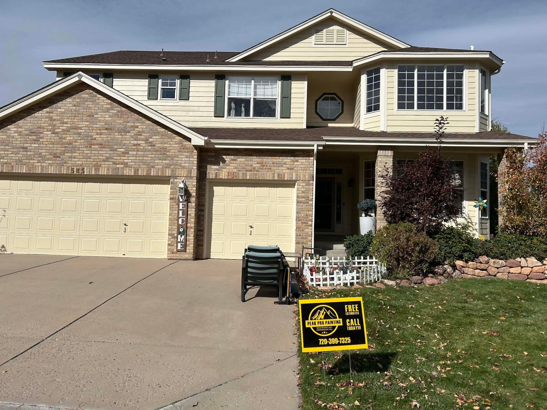 A two-story suburban home with brick and tan siding, a three-car garage, and a yard sign in the front lawn.