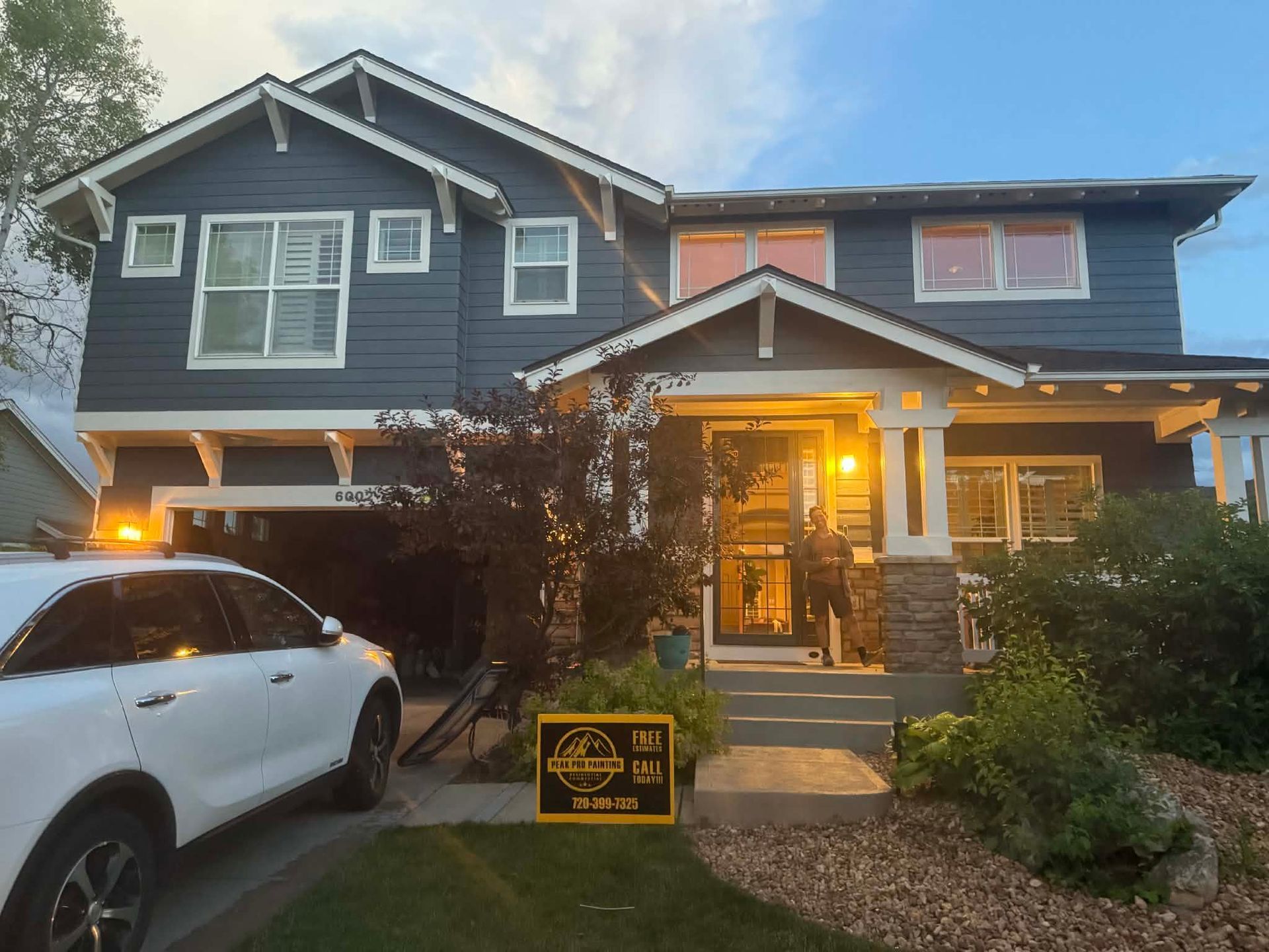 A blue two-story house with a white garage, a front porch with a glowing light, and a white car parked in the driveway.