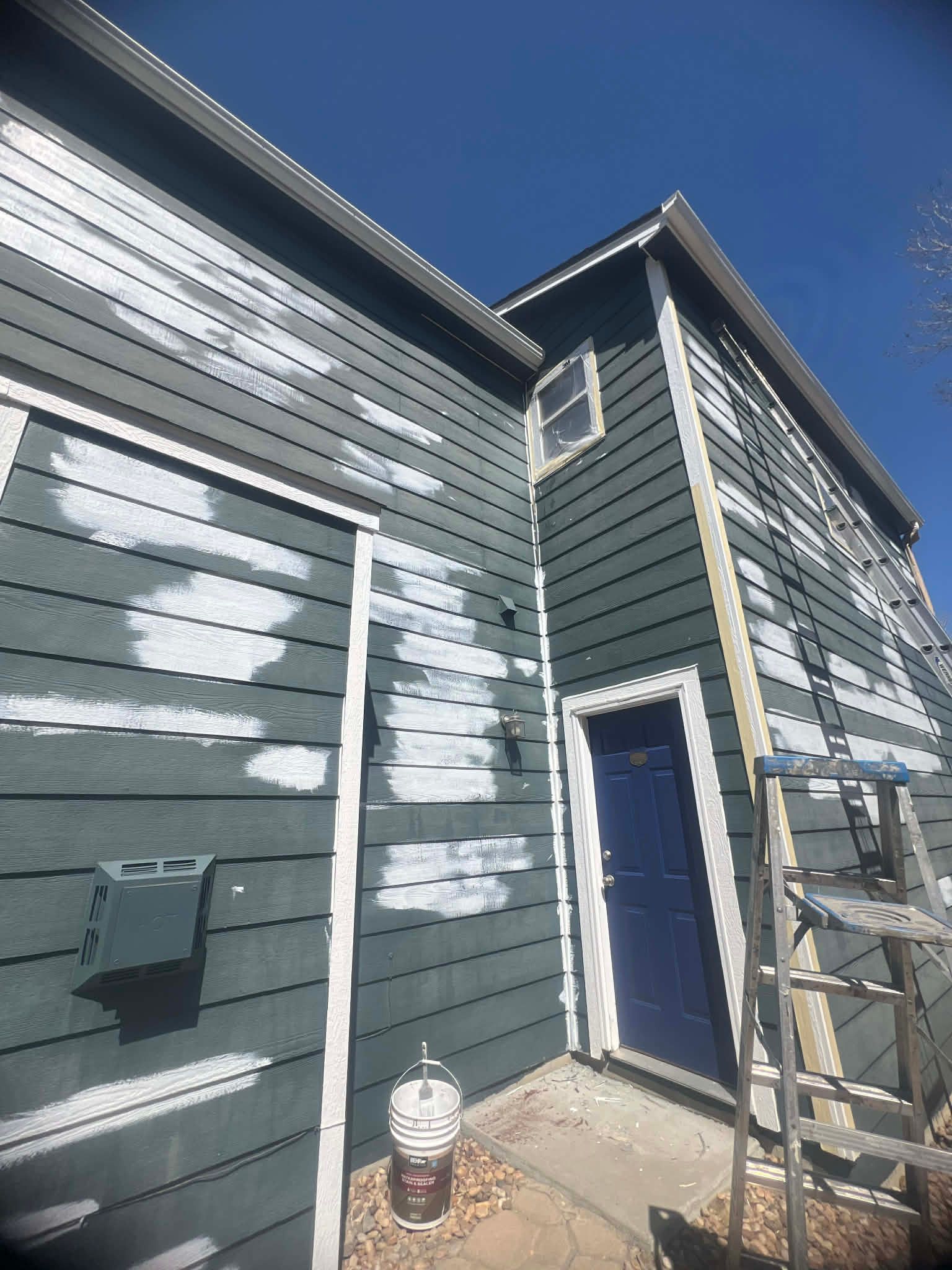 Exterior wall of a dark-sided house under repair with white primer patches and a ladder leaning against it.