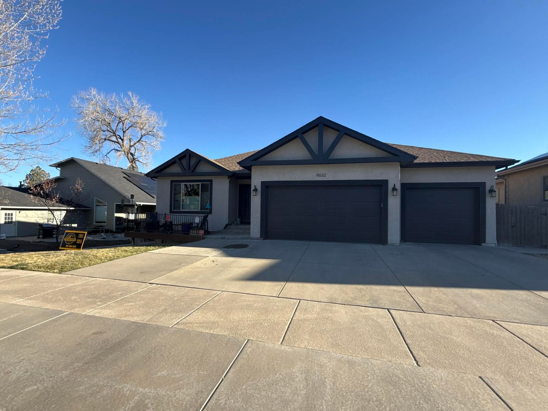 A one-story suburban home with a large concrete driveway, gray garage doors, and a clear blue sky.