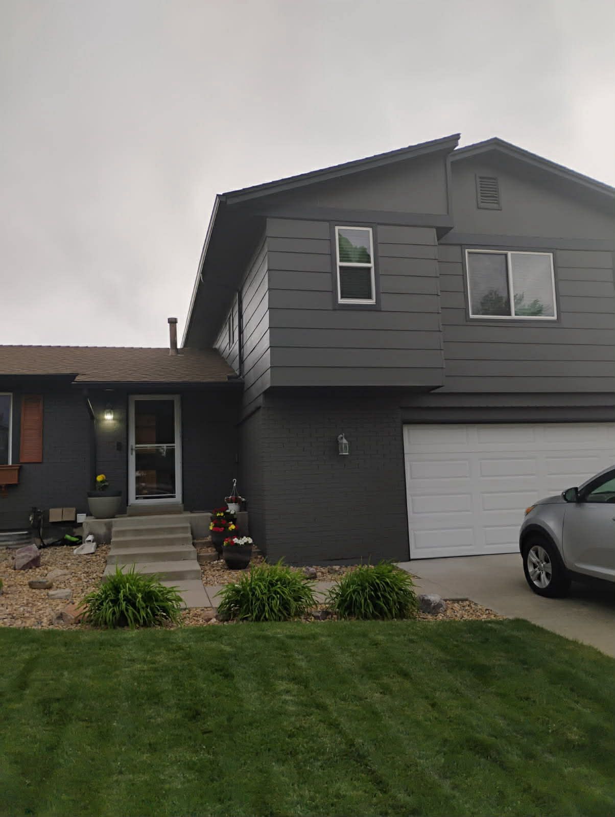 A two-story dark gray house with a white garage door, concrete steps to the entrance, and a green lawn in the foreground.