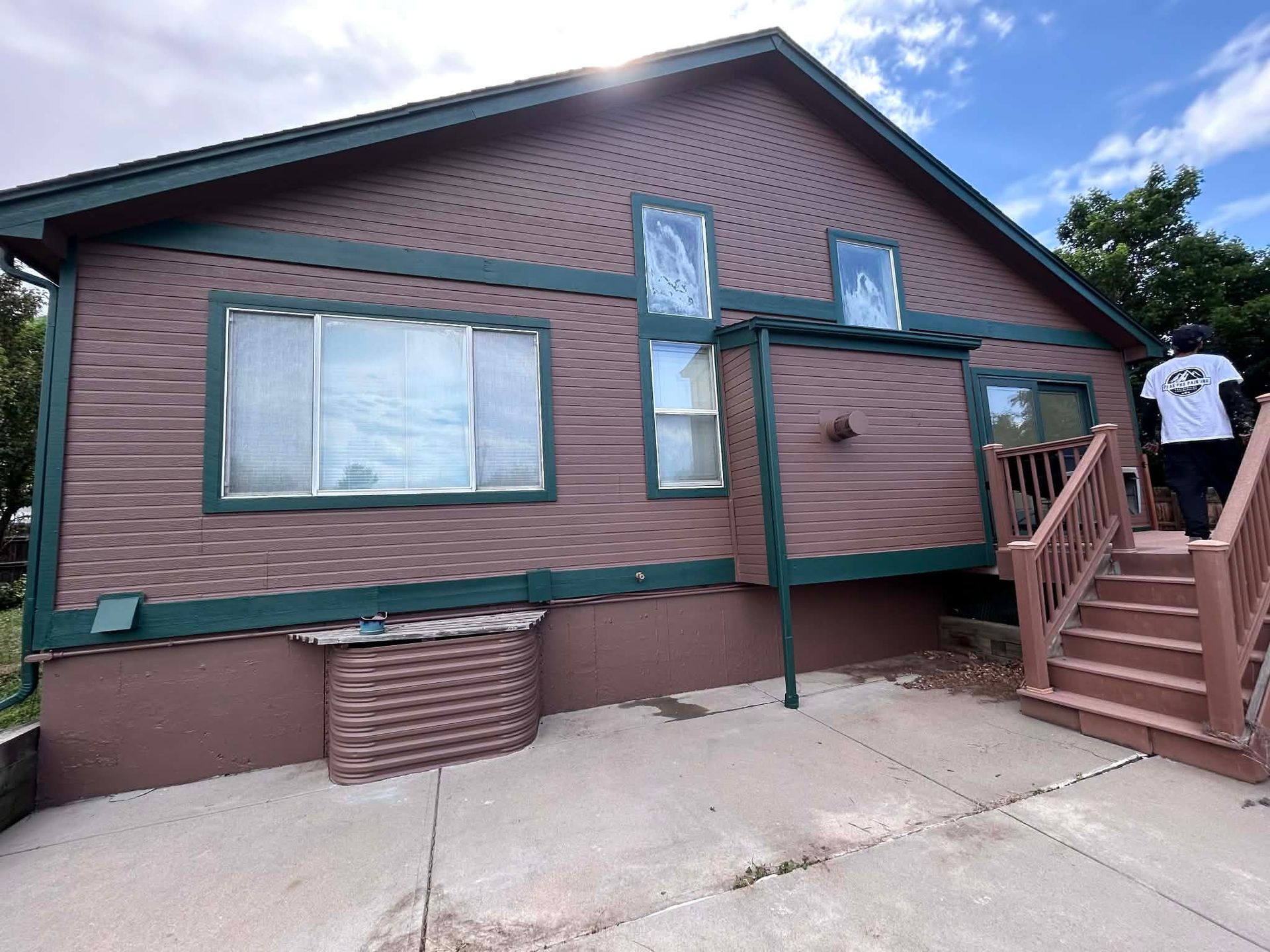 Brown house with green trim, a patio, a window well, and wooden stairs with a person walking up them on a sunny day.