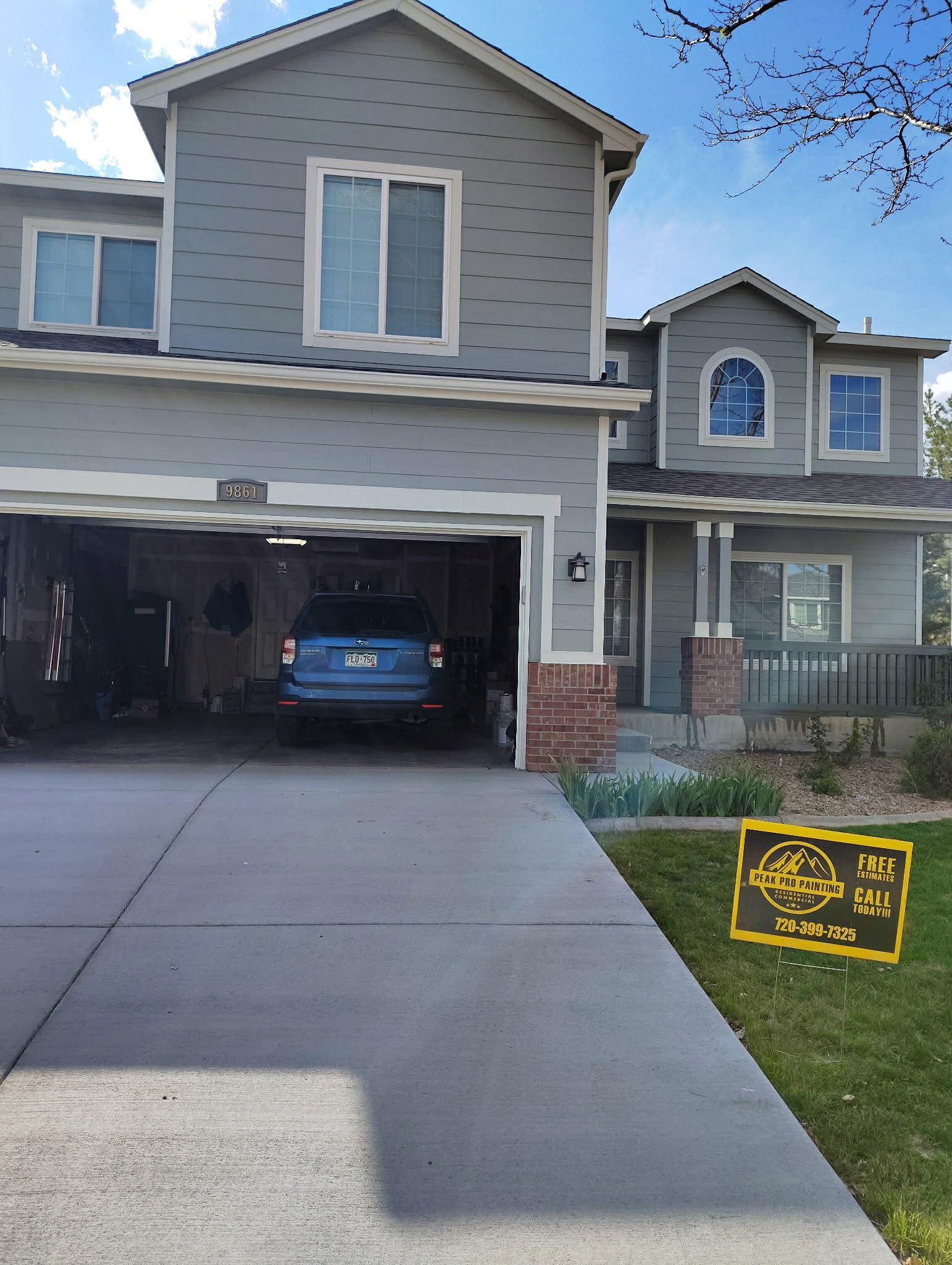 A two-story grey suburban house with a brick base, a garage with a blue car, and a yard sign on the lawn.
