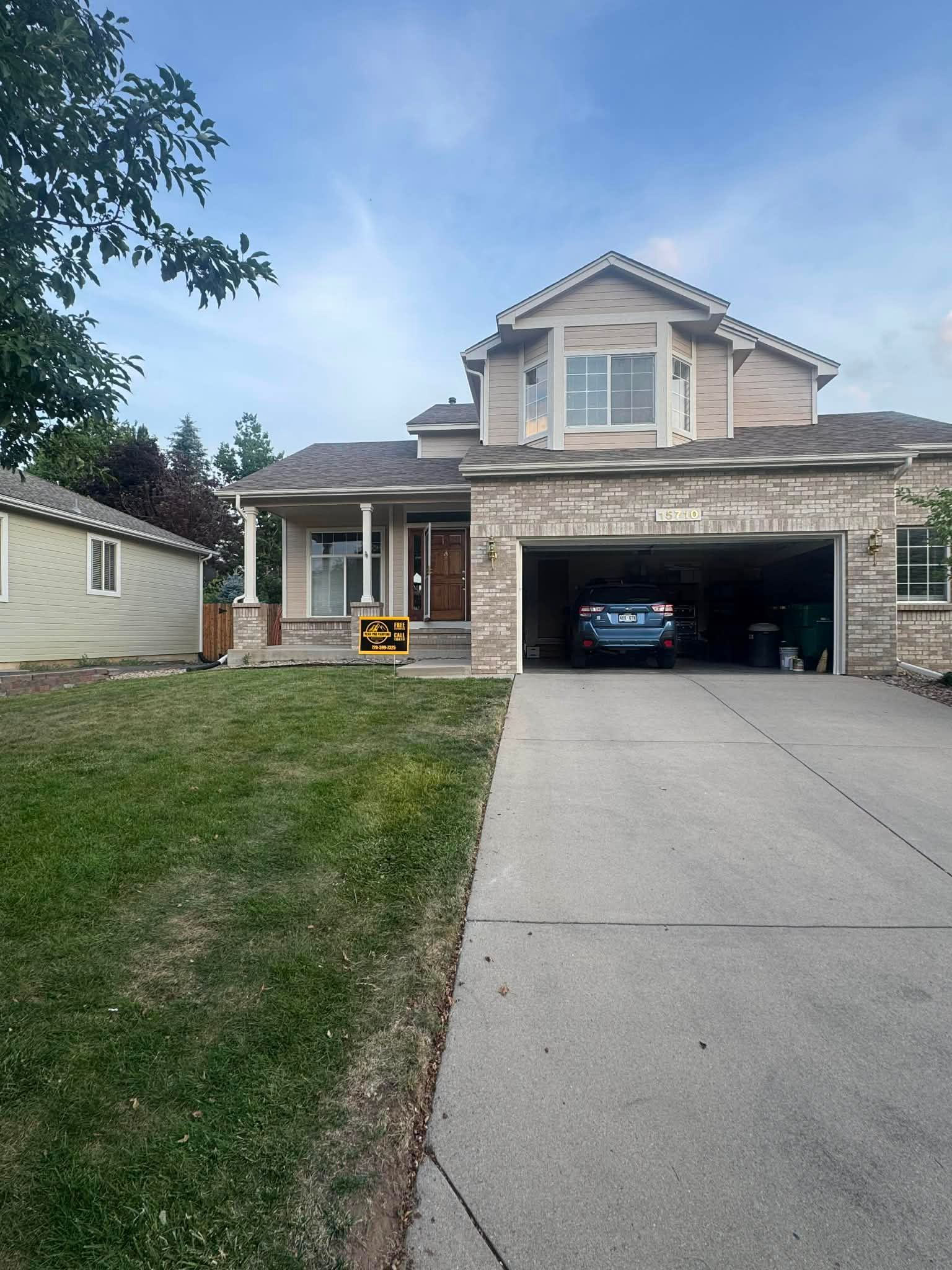 A tan two-story house with a brick facade and a two-car garage sits behind a concrete driveway and a grassy front lawn.