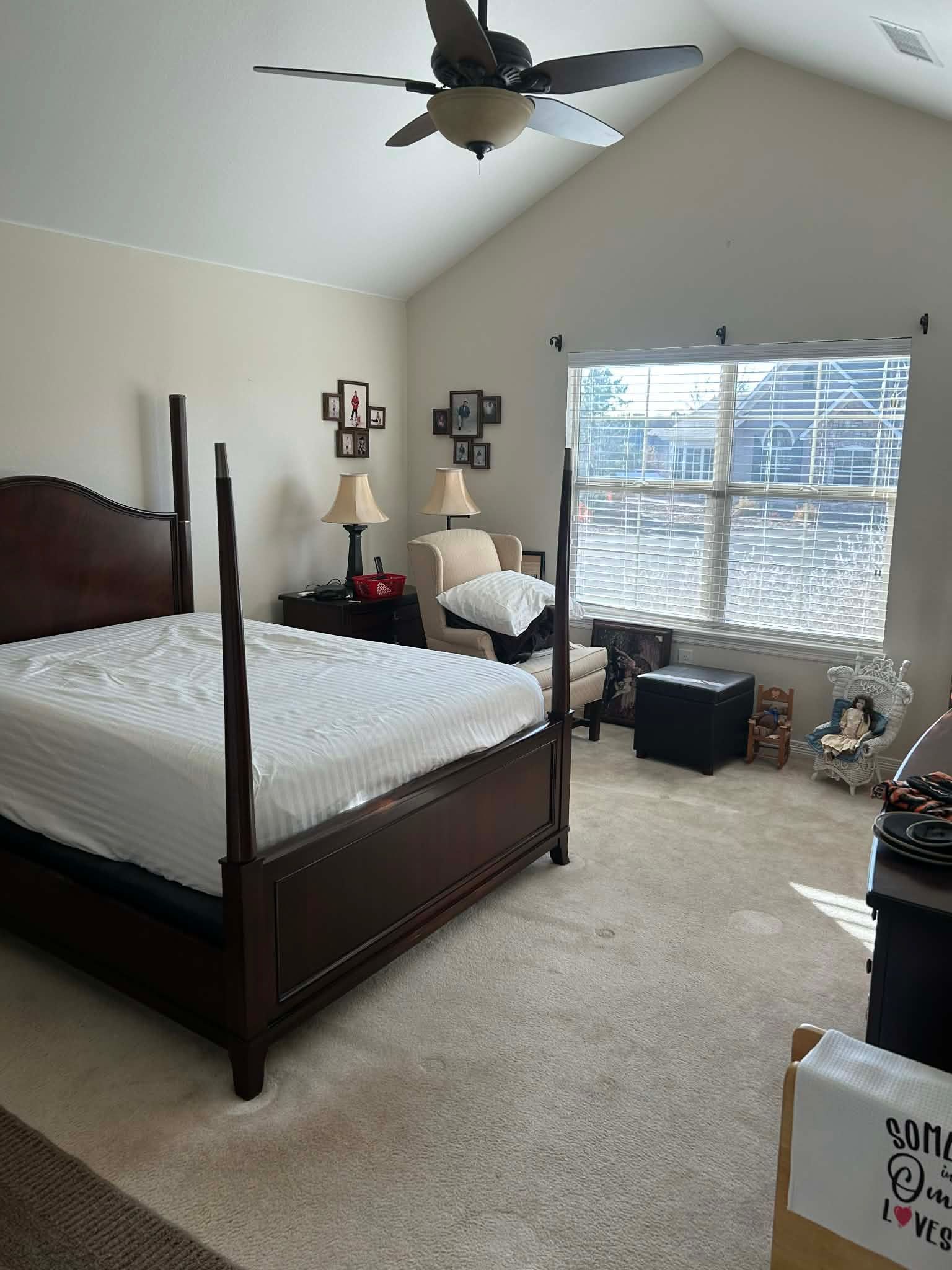A bedroom with a dark wood poster bed, beige walls, vaulted ceiling, and a large window next to an armchair.