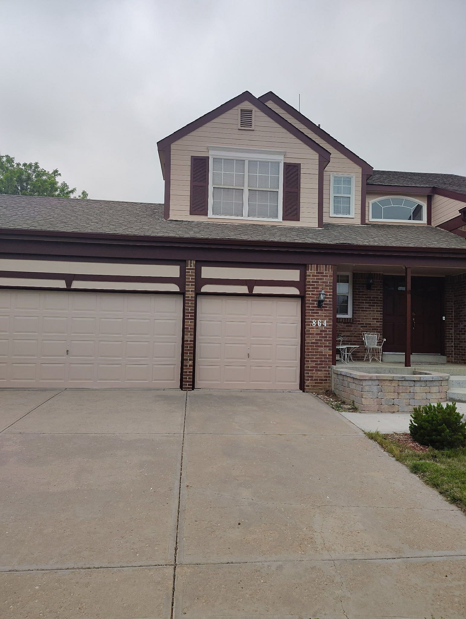 A two-story tan house with a dark brown trim, a large two-car garage, and a brick-accented entryway under a cloudy sky.