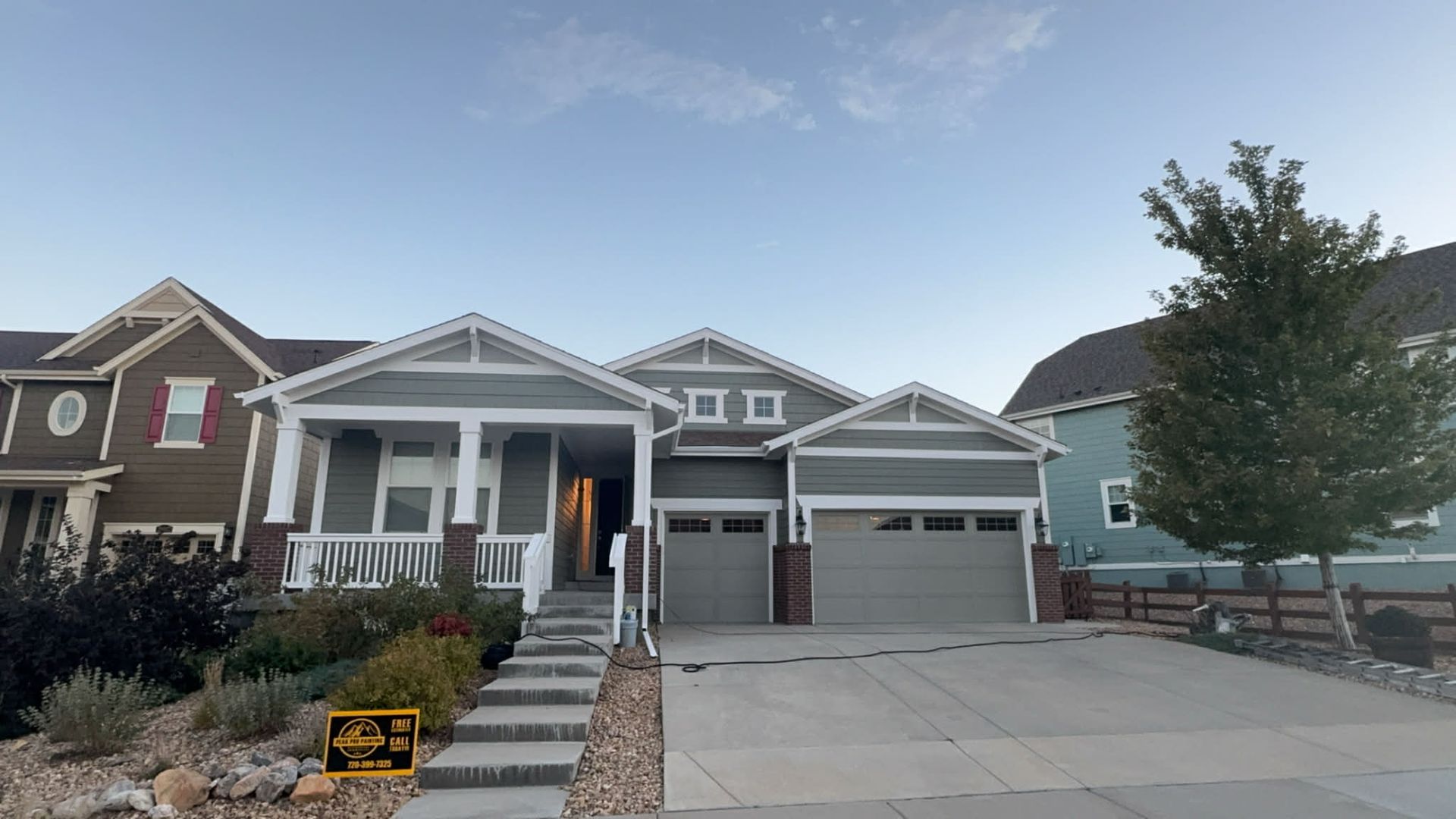 A modern gray house with a two-car garage, a front porch with white columns, and stone steps, flanked by other homes.