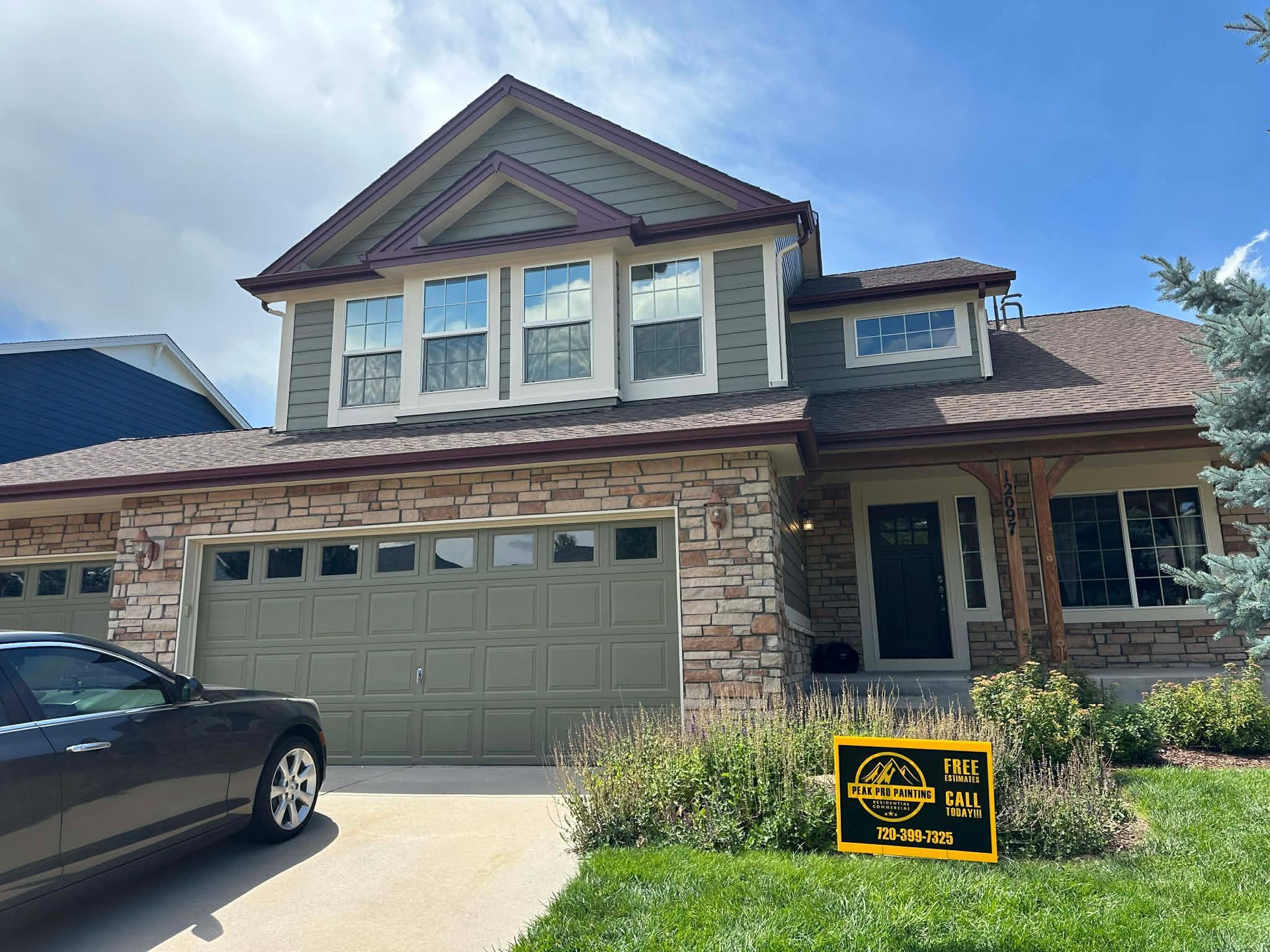 A two-story suburban home with stone and green siding, a two-car garage, and a real estate sign in the front yard.