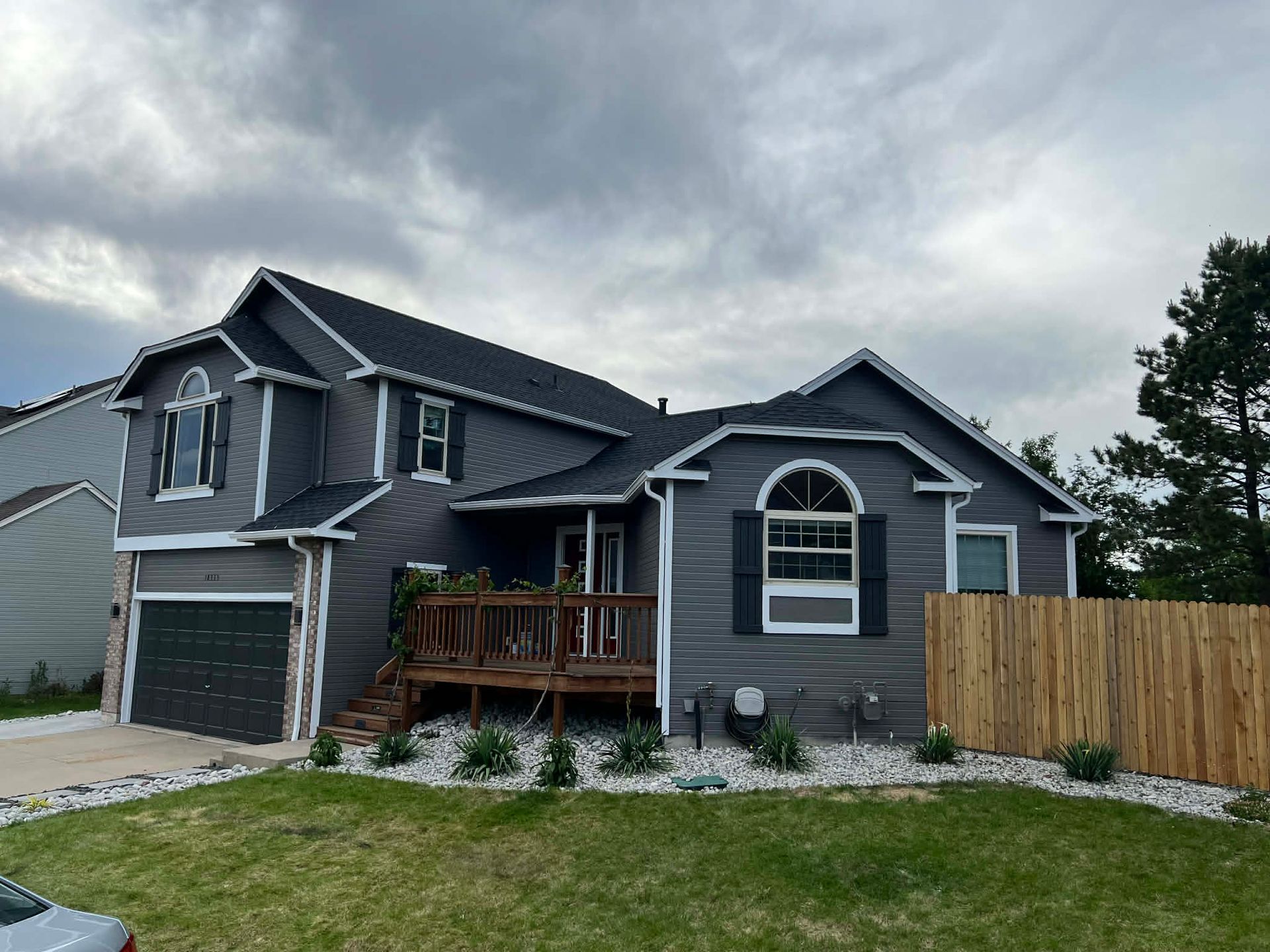A two-story, dark gray house with a wooden front deck, white trim, a dark garage, and a light wood fence under cloudy skies.