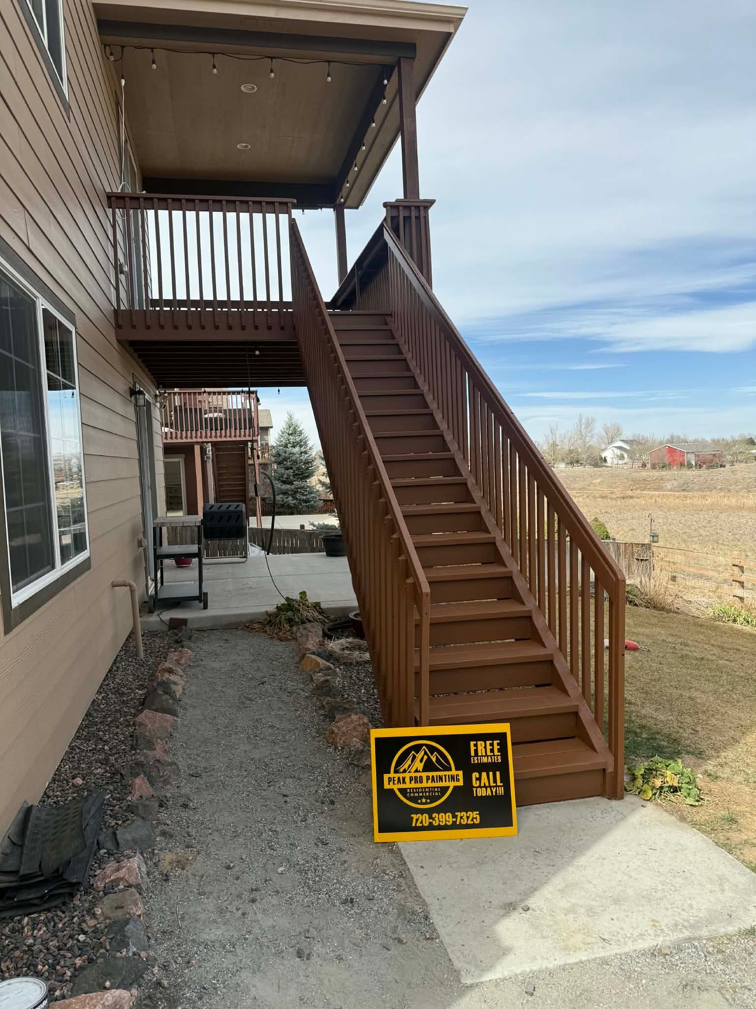 A brown wooden deck with stairs leads down to a concrete patio and a gravel walkway next to the side of a house.