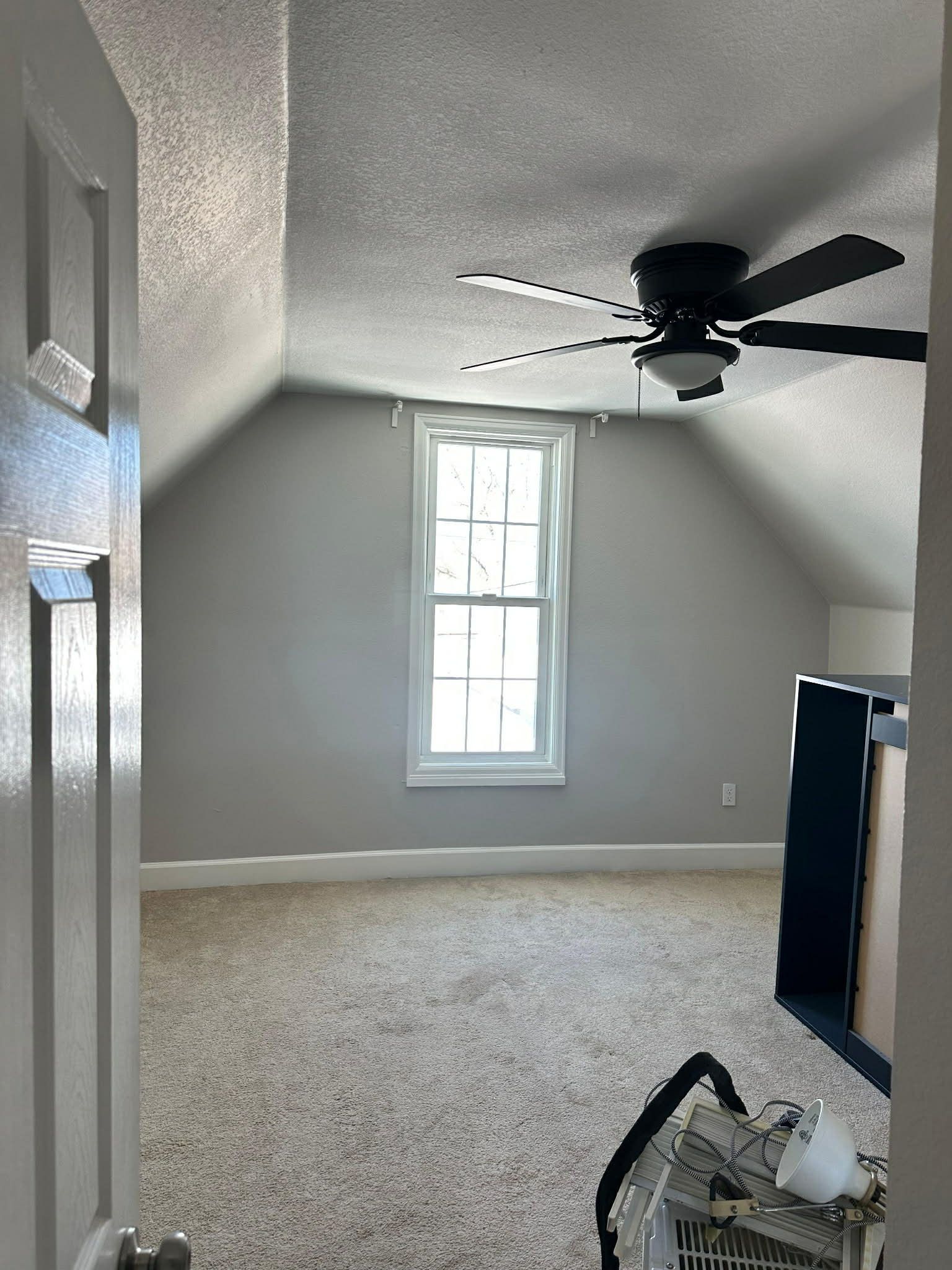 An empty attic room with gray walls, a window, tan carpet, and a ceiling fan, viewed from a doorway.
