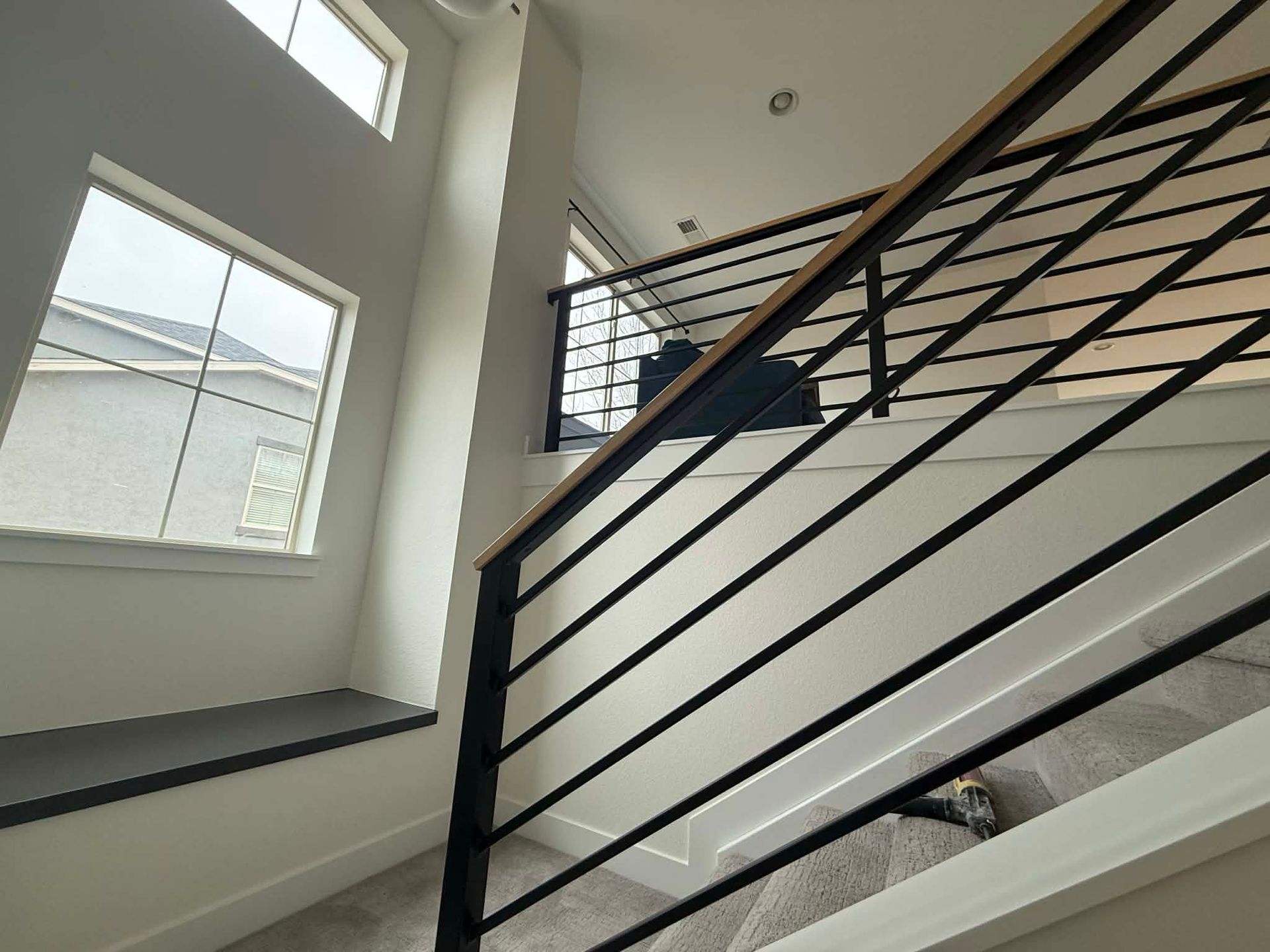 A wooden handrail with black horizontal metal balusters runs along a carpeted staircase next to a window seat.